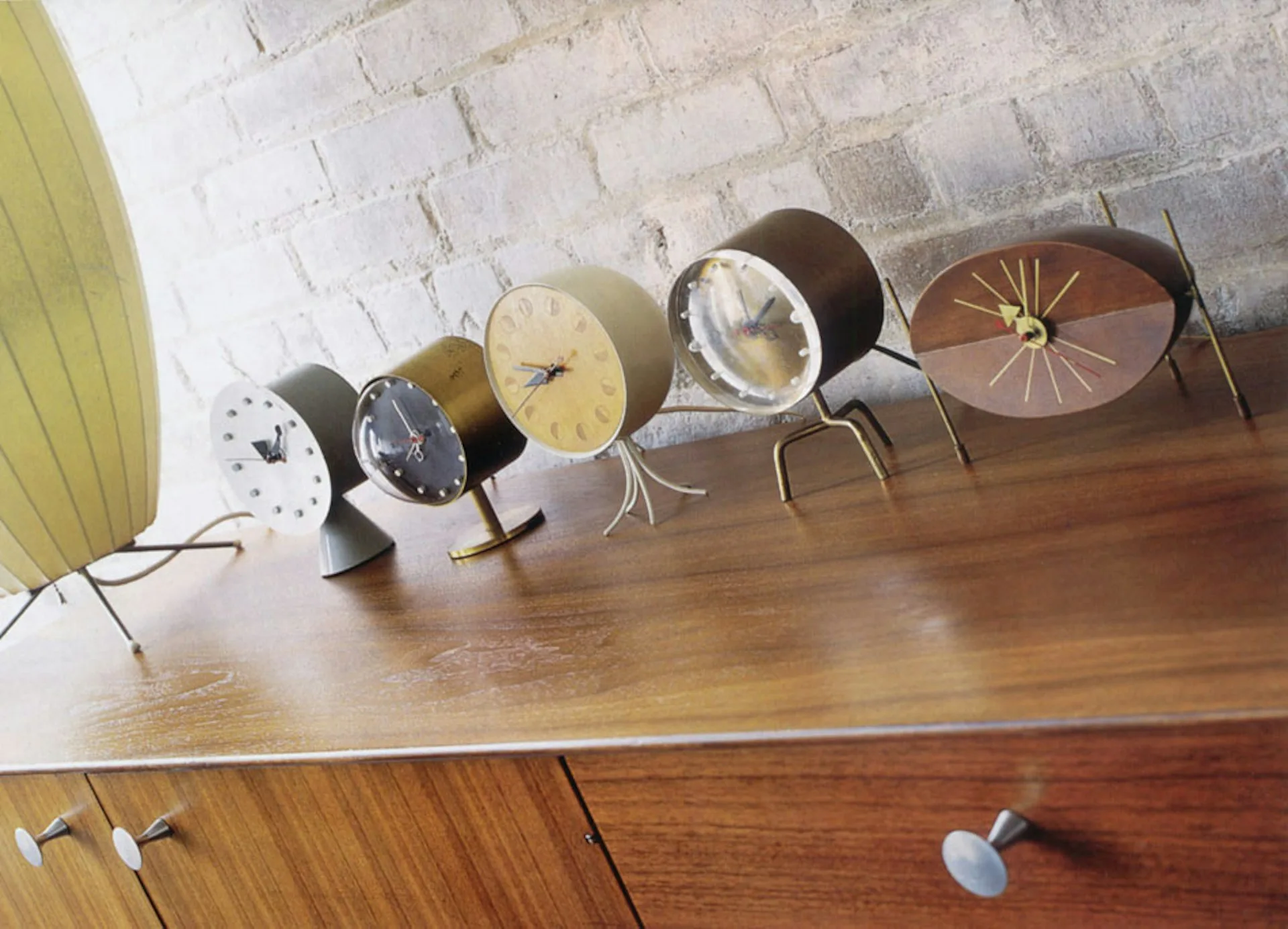 Collection of mid-century modern table lamps with various dial faces and metal bases arranged on a wooden surface against a white brick wall by James Wagman Architect, LLC.