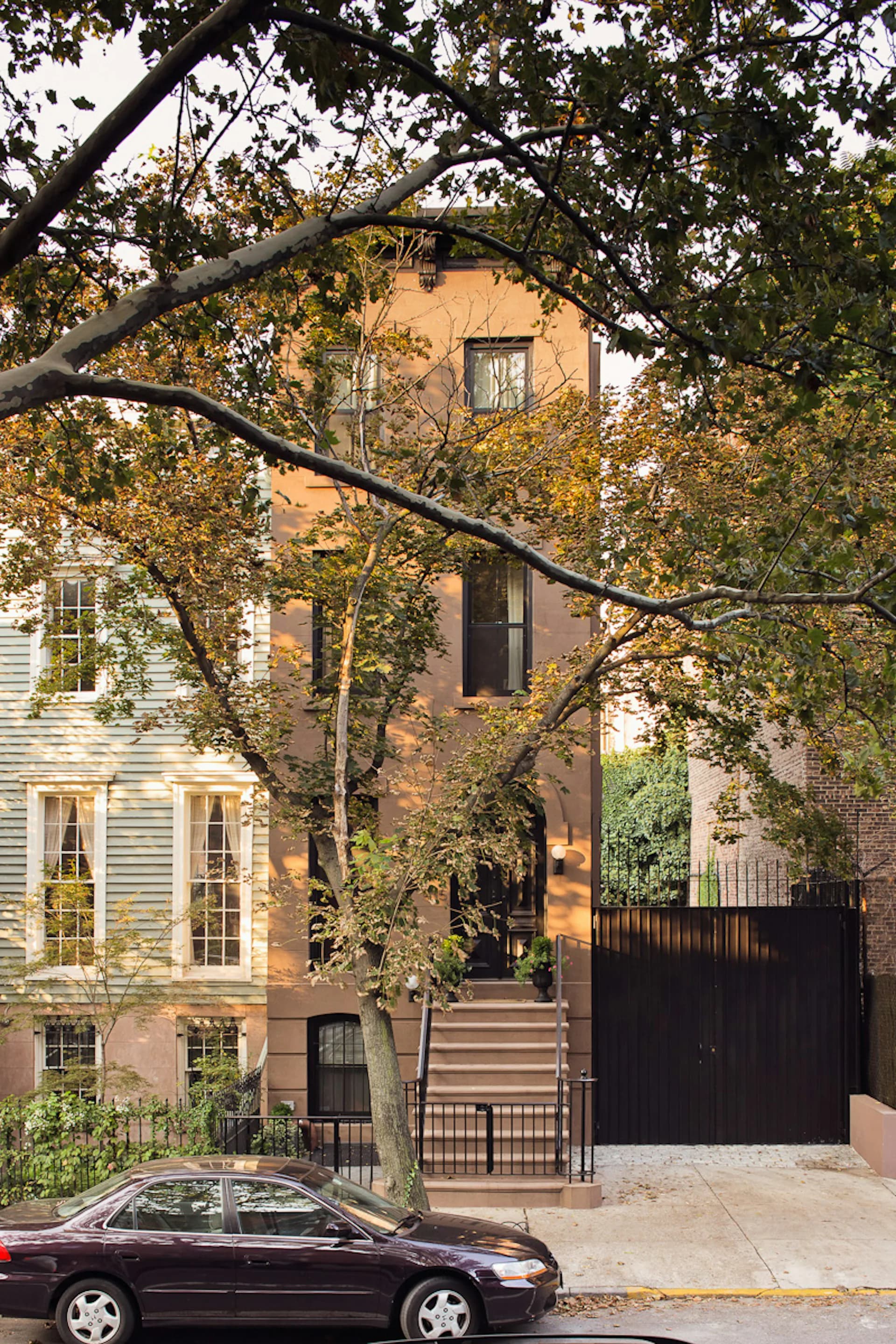 Warm terracotta and brick residential building with modern black gate and metal railings, framed by mature tree branches in autumn, by Janko Rasic Architects, PLLC.