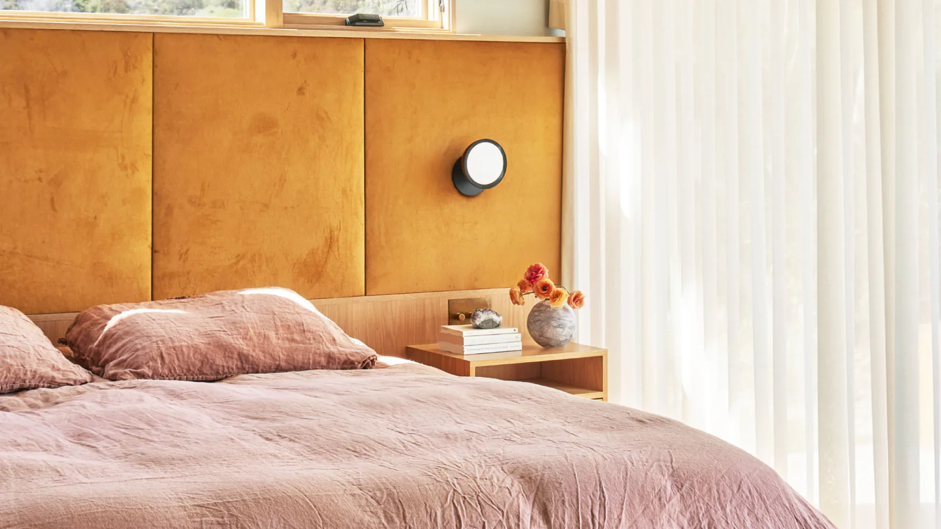 Warm bedroom with cognac leather headboard, blush pink bedding, integrated nightstand, and circular wall sconce by Jason Miller Studio.