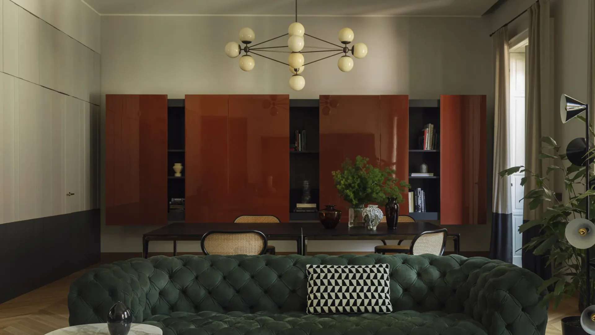 Modern living room interior featuring a tufted green velvet sofa, rust-red cabinetry with black shelving, geometric patterned accent chair, and sculptural brass pendant lighting by Jason Miller Studio.