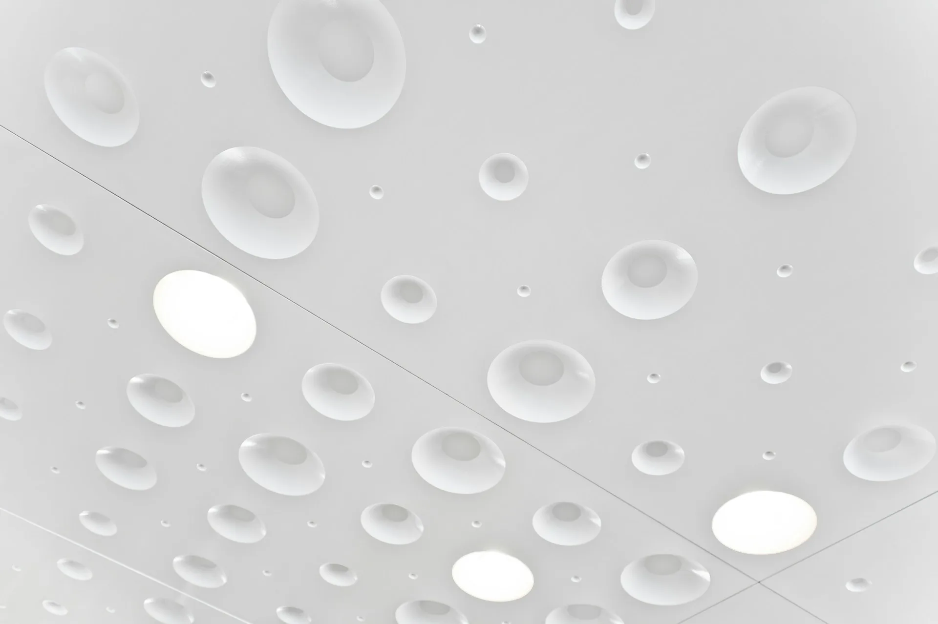 Overhead view of a geometric ceiling with recessed circular and elliptical light fixtures in varying sizes against a pale gray surface by Joeb Moore & Partners.