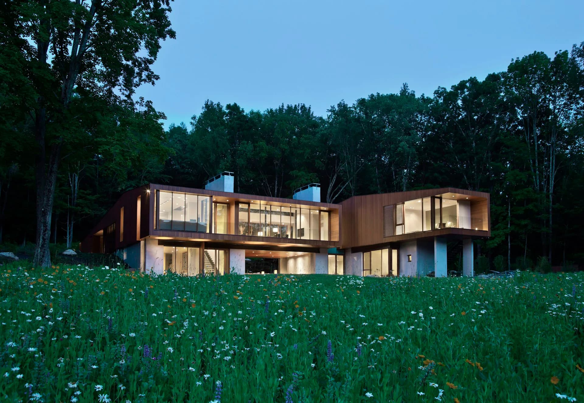 Modern residential architecture with curved wood and glass forms set in a wildflower meadow at dusk, illuminated interiors visible, surrounded by forest by Joeb Moore & Partners.