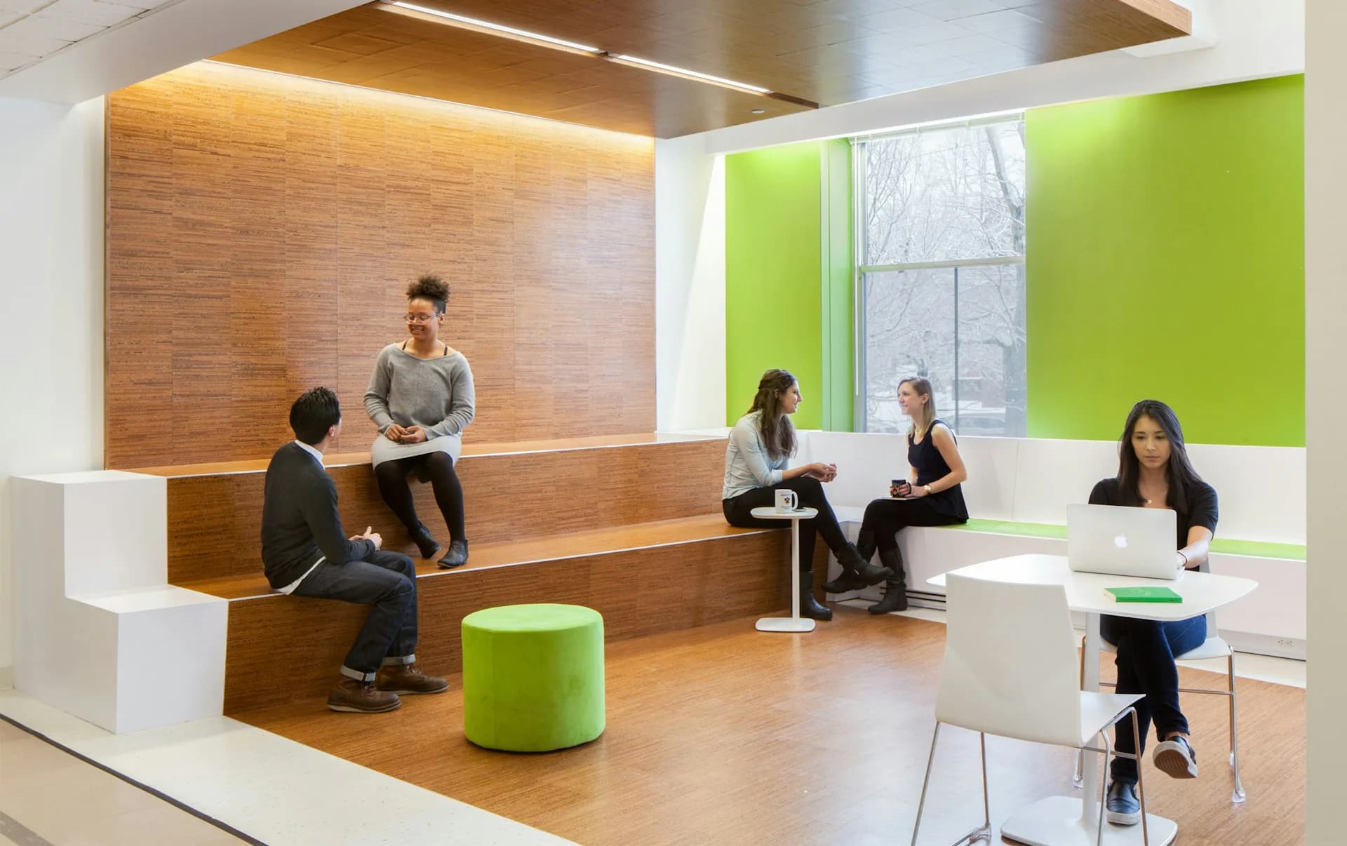 Modern office breakroom with warm wood paneling, lime-green accents, and open collaborative seating areas with natural light by Joel Sanders Architect.