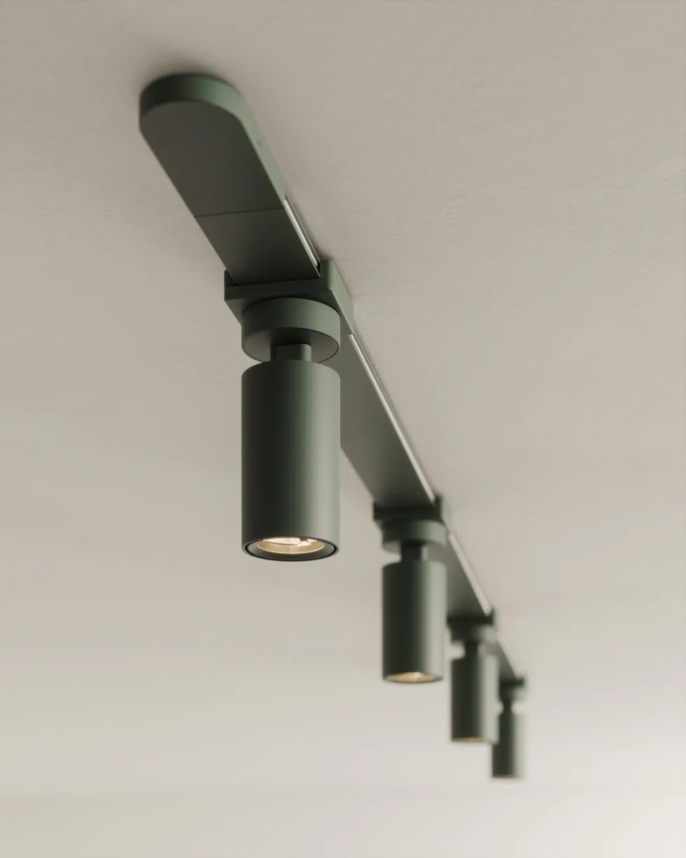 Angled ceiling-mounted track lighting fixture with multiple cylindrical green metal heads emitting warm light, photographed against a neutral ceiling by Juniper Design.