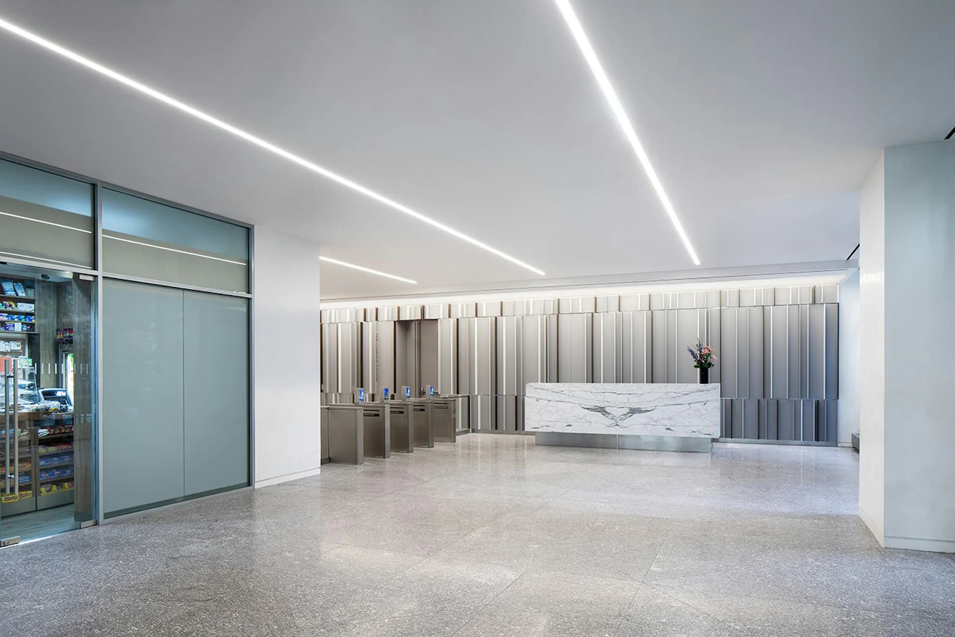 Modern commercial lobby featuring polished concrete floors, linear ceiling lights, marble-clad reception desk, and vertical metal panel wall detail by Kahn Architecture & Design, PC.