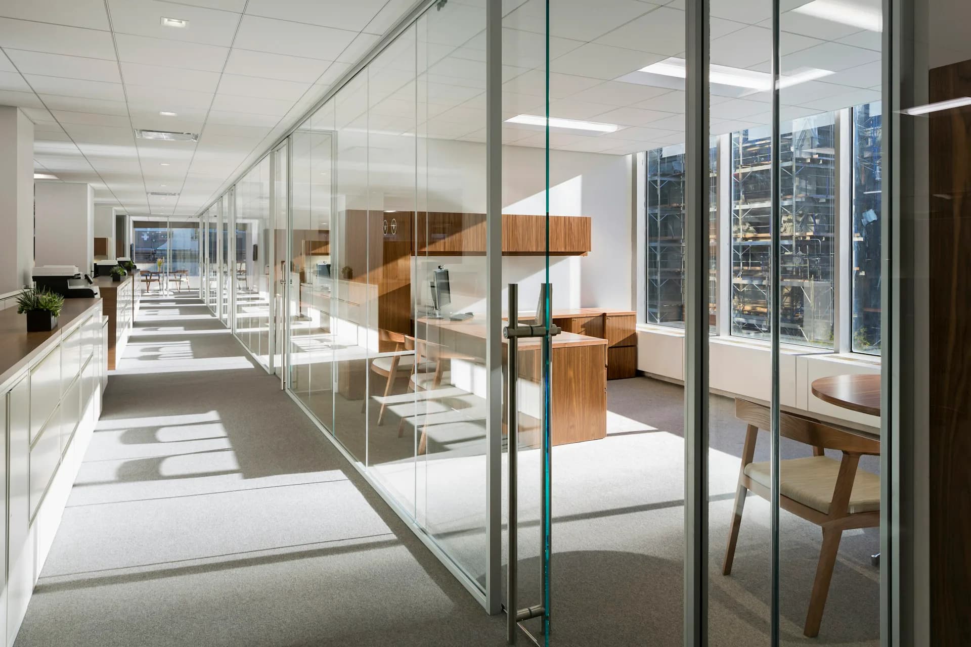 Modern office corridor with floor-to-ceiling glass partitions, warm wood cabinetry, and panoramic city skyline views, designed by Kahn Architecture & Design, PC.