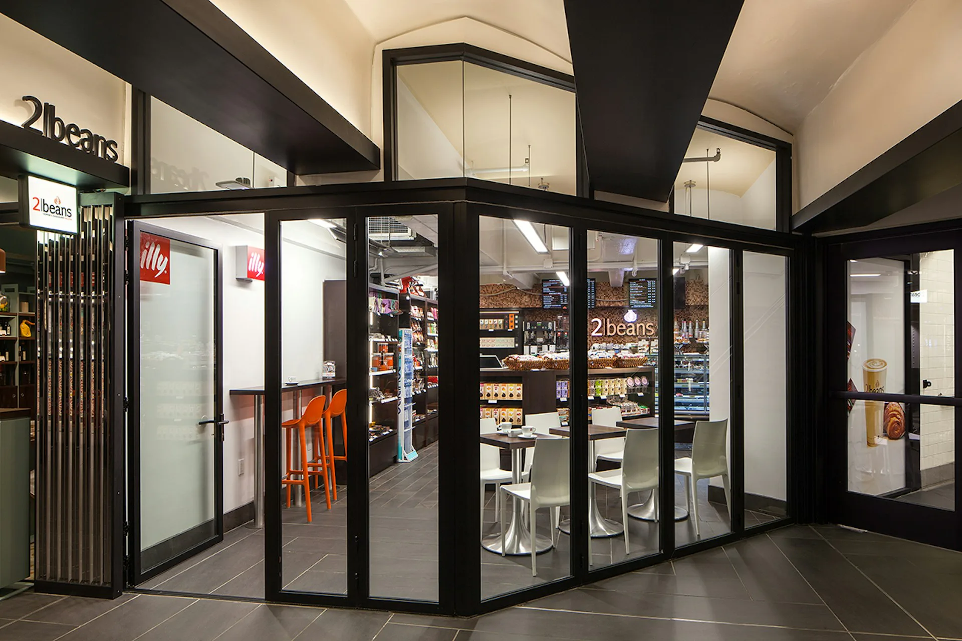 Corner retail storefront with black framing, glass doors and windows, and illuminated interior displaying products and shelving, designed by Kahn Architecture & Design, PC.