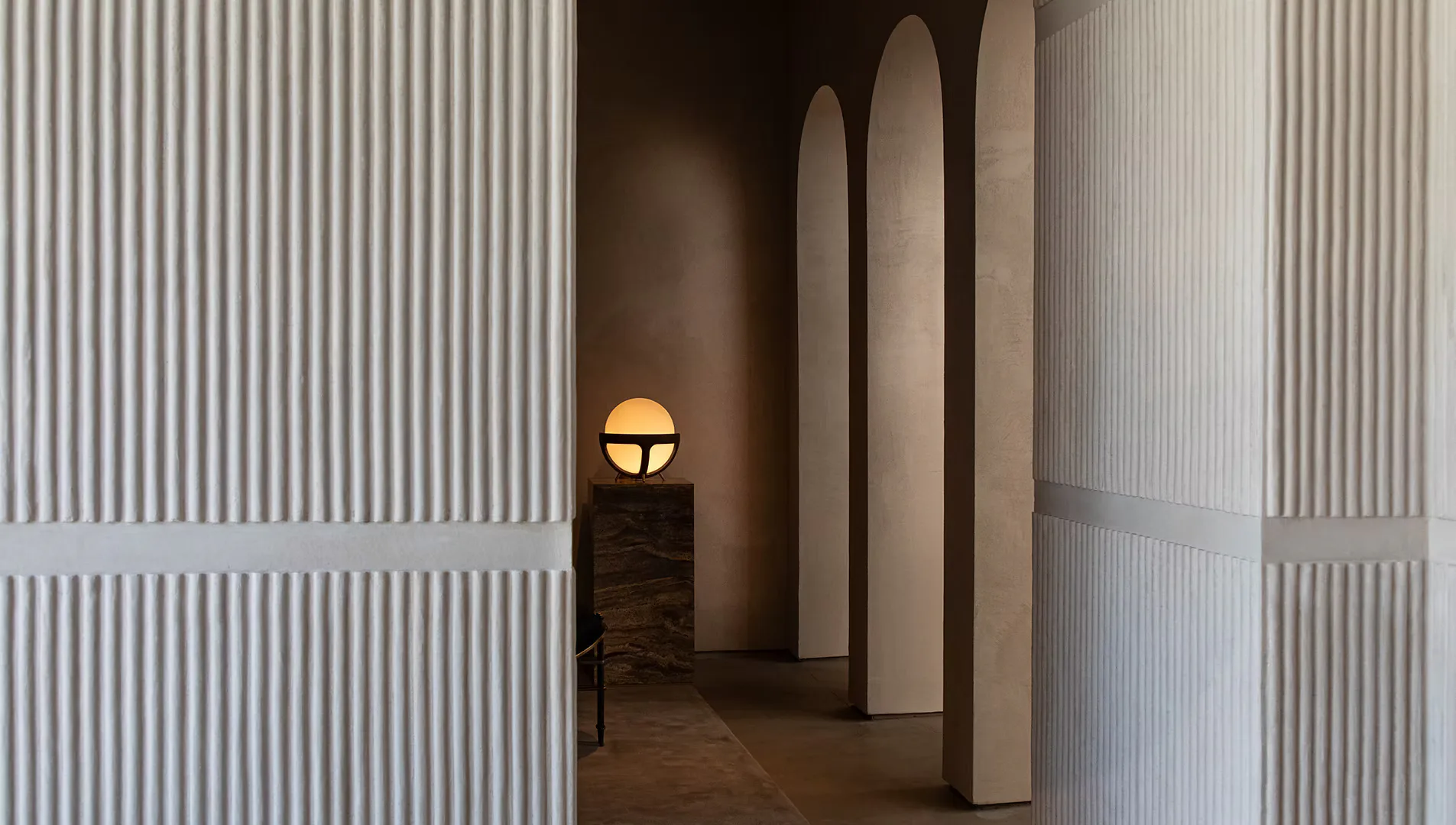 Minimalist corridor with fluted white wall panels, tall arched openings, warm accent lighting, and dark flooring by Kamp Studios.