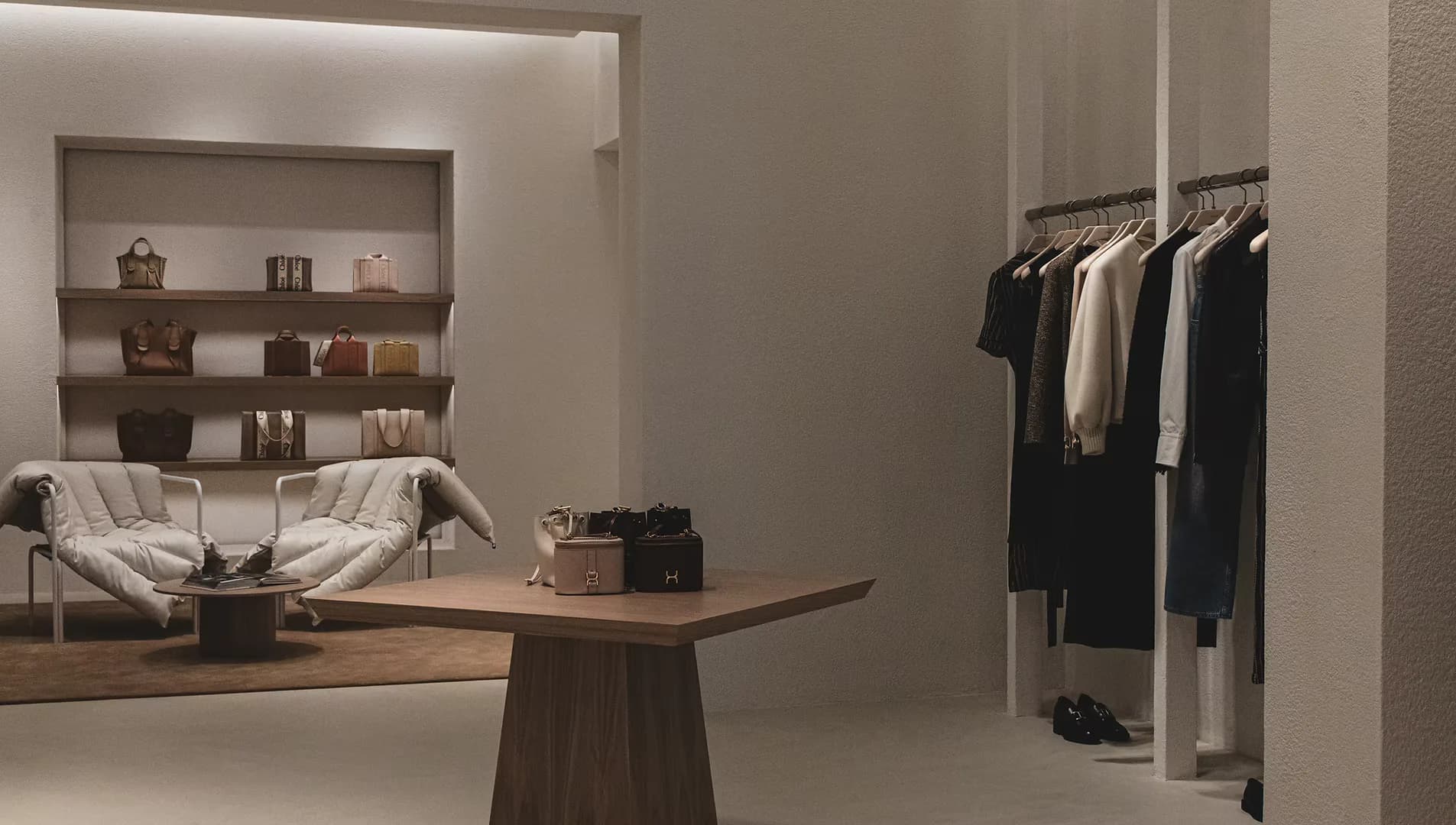 Modern retail interior with neutral palette featuring wooden display table, wall-mounted shelving, clothing rack, and cream papasan chair by Kamp Studios.