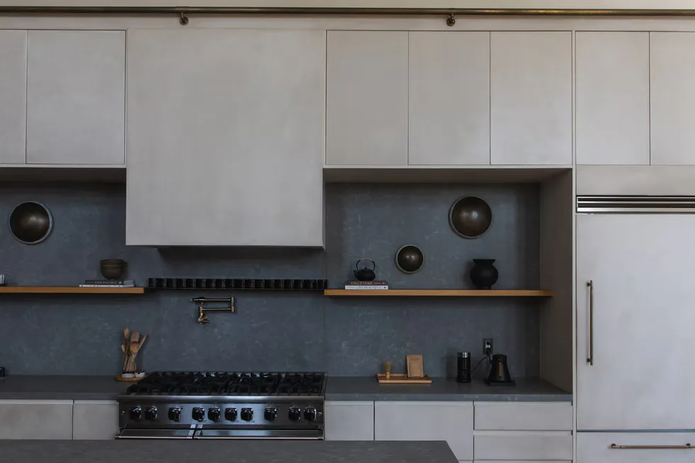 Modern kitchen with white upper cabinets, dark gray tile backsplash, stainless steel appliances, and open shelving with decorative objects by Kamp Studios.