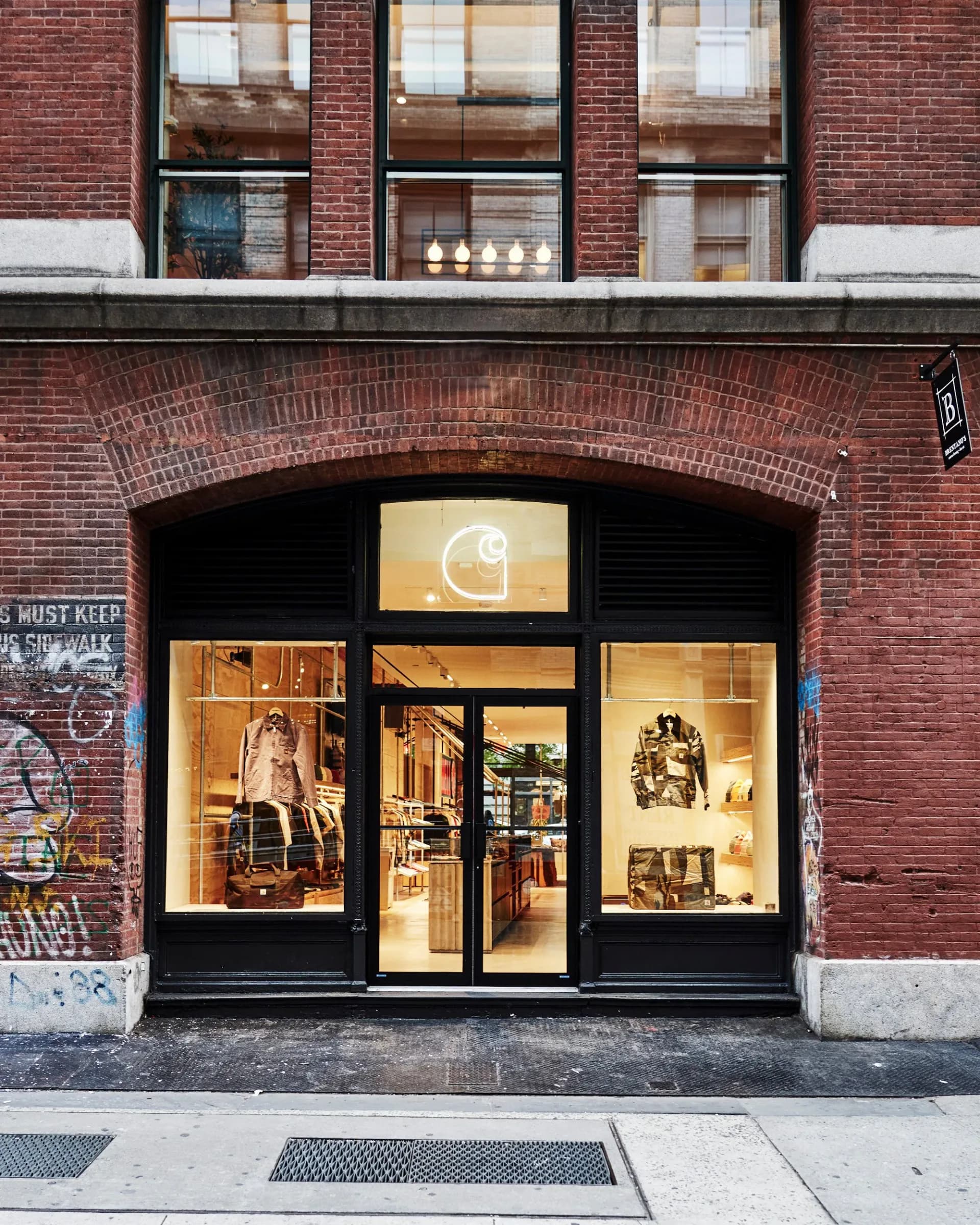 Storefront featuring a black-framed glass entry with warm wood and stone interior finishes, arched brick facade above, and illuminated windows displaying retail goods by Katz Architecture.