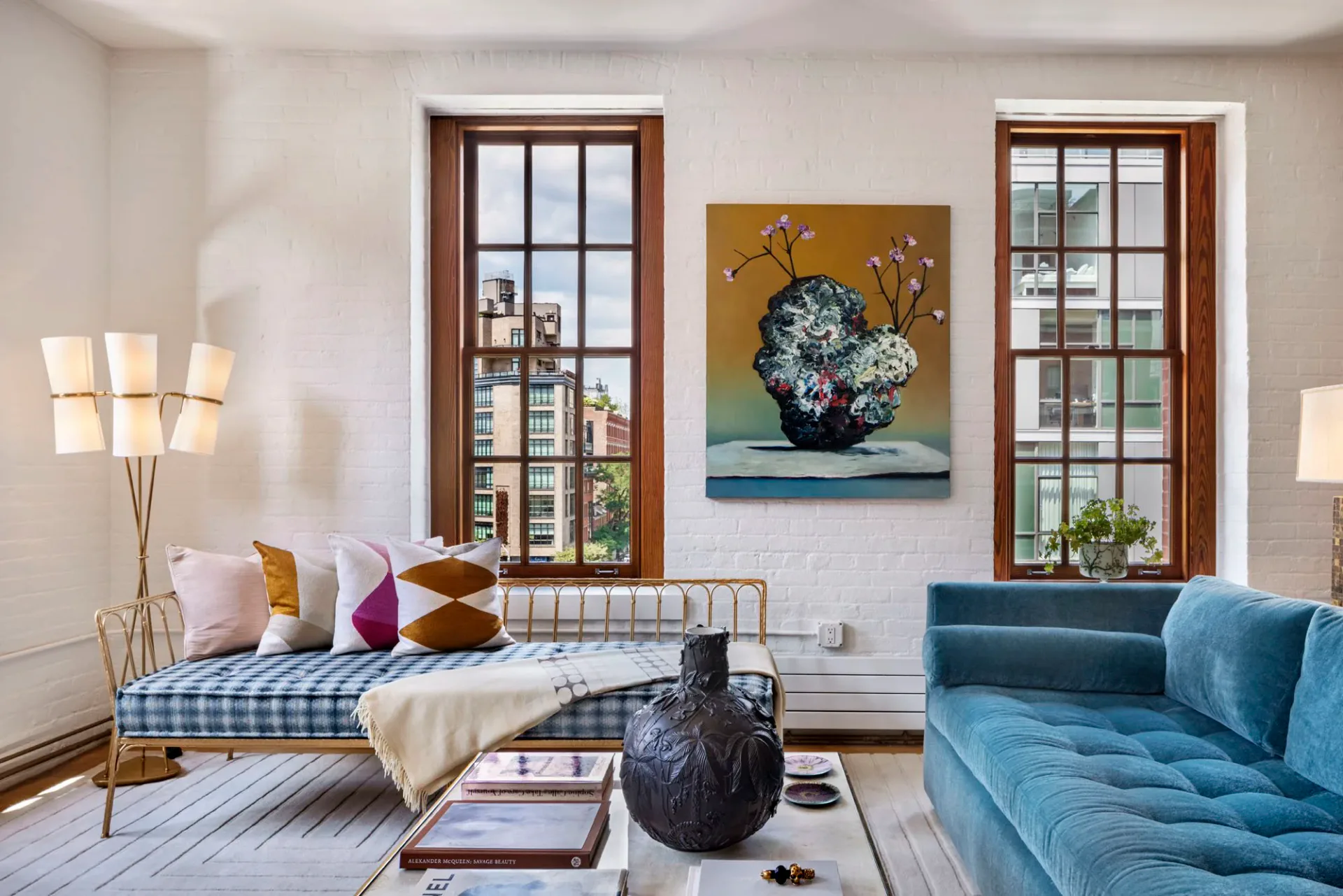Bright residential bedroom with brass bed frame, blue upholstered sofa, large windows with wood frames, gallery wall artwork, and layered lighting design by Katz Architecture.