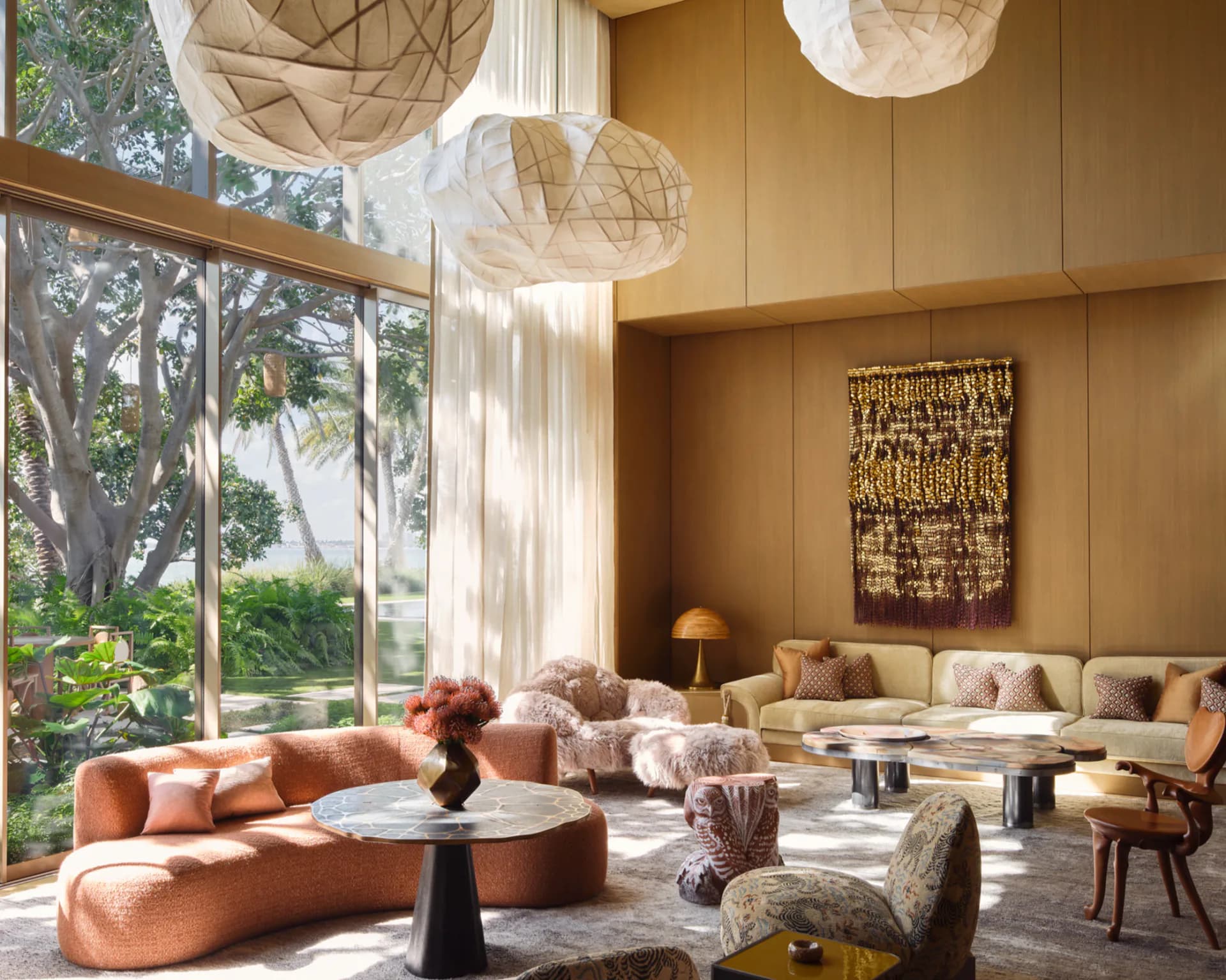 Warm-toned luxury living room with curved coral sofa, geometric pendant lights, and floor-to-ceiling windows overlooking trees, designed by Kelly Behun Studio.