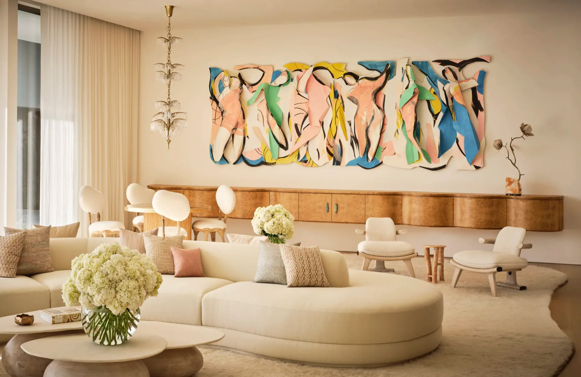 Modern living room with curved cream sofa, wooden sideboard, colorful abstract artwork, and warm neutral palette by Kelly Behun Studio.