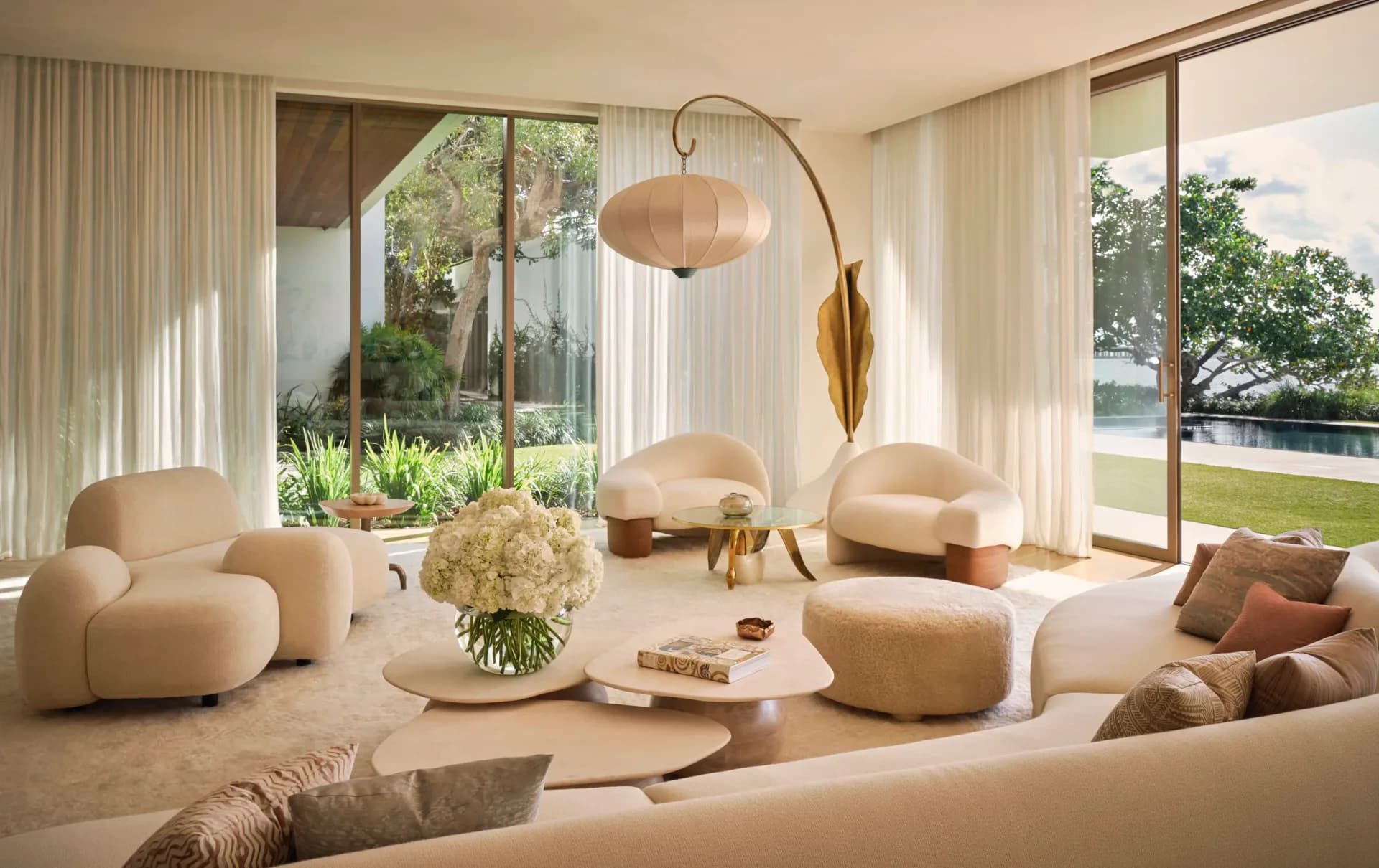 A minimalist living room with curved cream furniture, arched floor lamp, and floor-to-ceiling windows overlooking green landscape by Kelly Behun Studio.