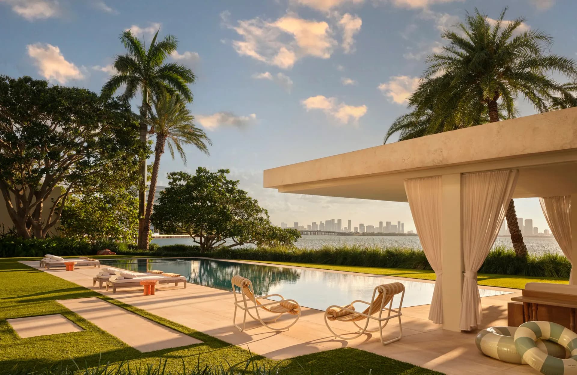 Waterfront residential landscape with infinity pool, modern pergola, palm trees, and Miami skyline view by Kelly Behun Studio.