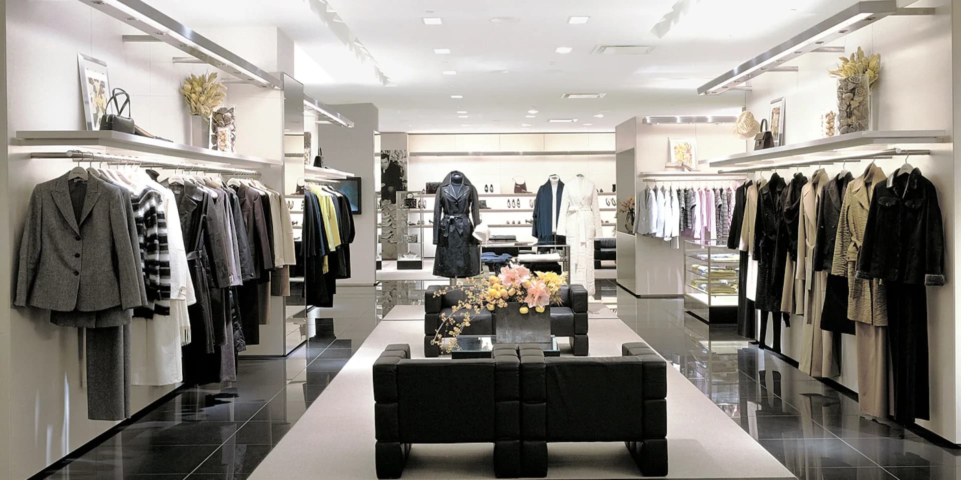 Modern luxury retail clothing showroom with black polished flooring, white walls, clothing racks on both sides, central black display table with floral arrangement, and recessed ceiling lighting by Kenne Shepherd Interior Design Architecture PLLC.