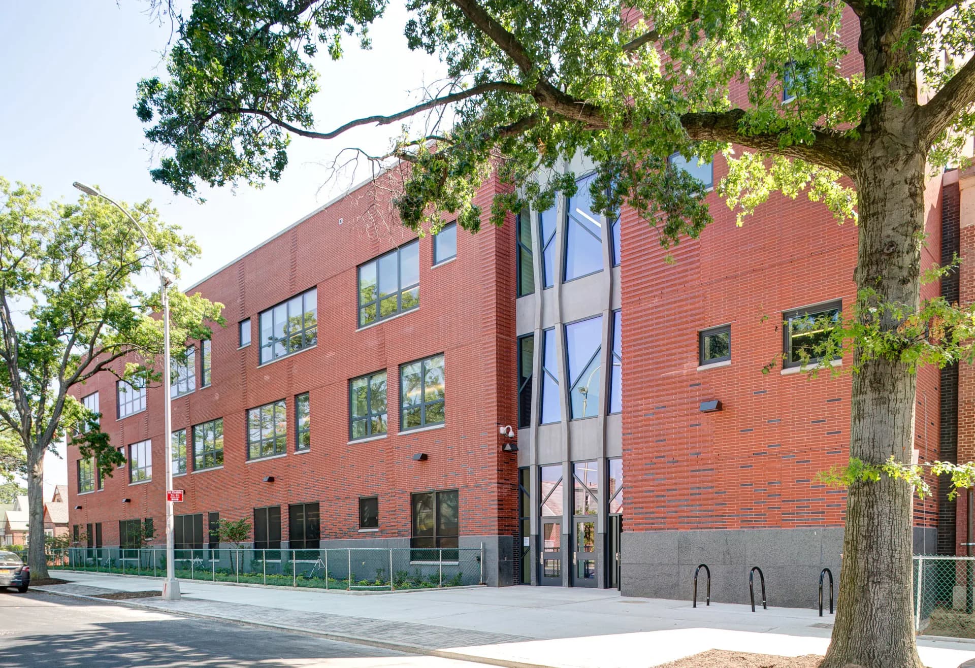 Red brick contemporary building with large glass entrance pavilion, surrounded by mature trees, designed by Kenny & Khan Architects PC.