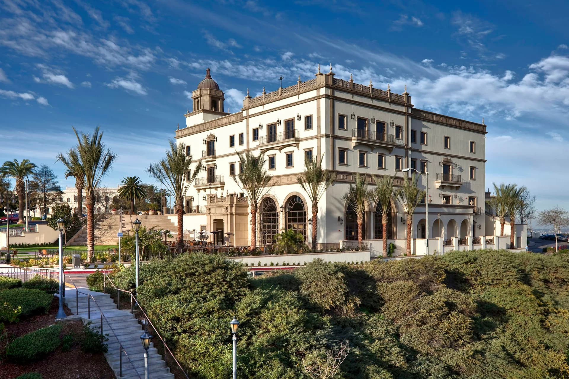 Elegant neoclassical palace with cream stone facade, arched colonnade, and dome, surrounded by manicured hedges and palm trees by Kevin Hom Architects.