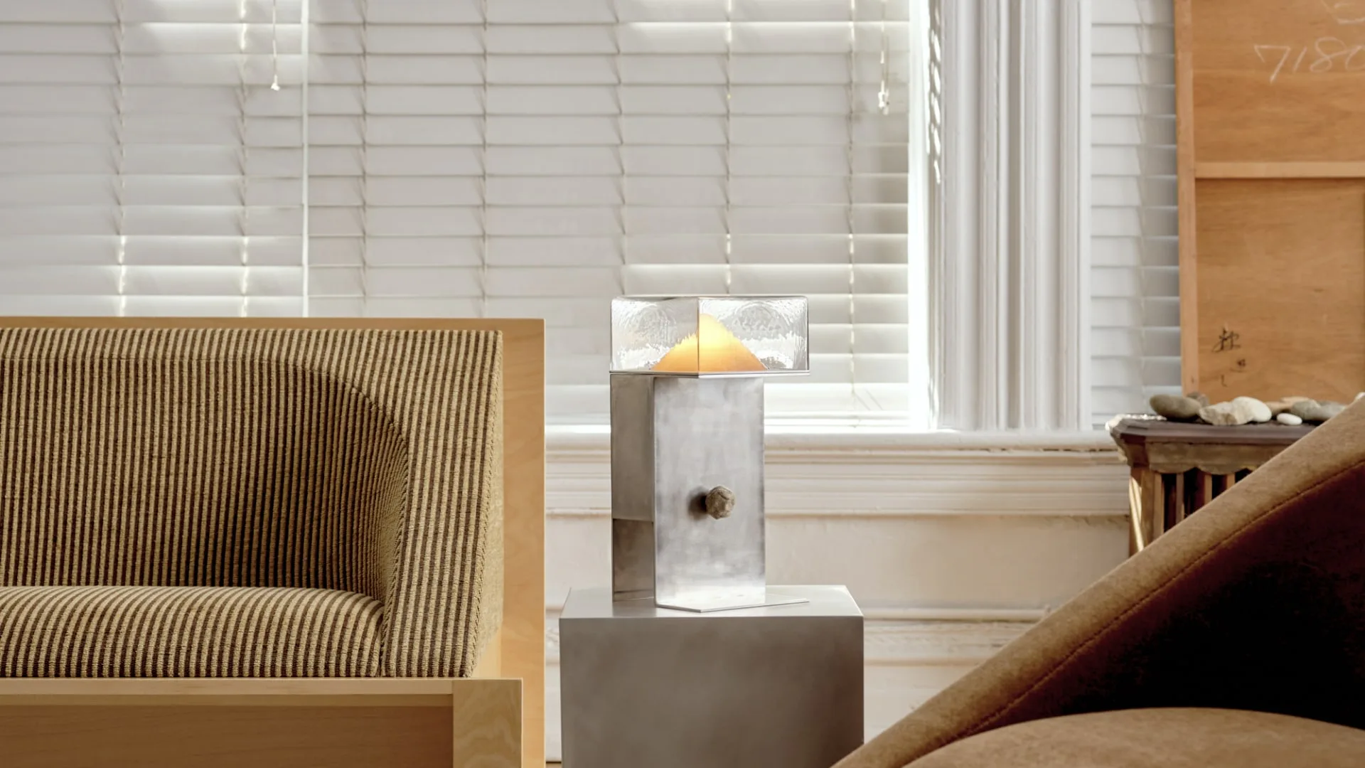 Minimalist interior with striped beige sofa, concrete side table with candle, and natural light from shuttered windows by Known Work.