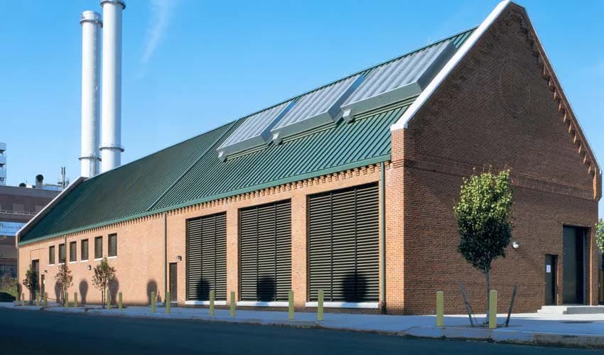 Contemporary brick industrial building with green metal roof and large glass windows, featuring white cylindrical smokestacks, designed by Koenen Associates.