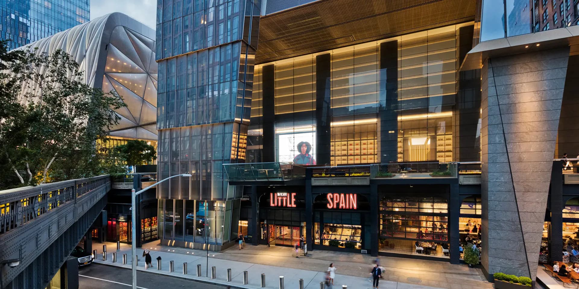 Modern mixed-use building facade with illuminated storefront featuring 'Little Spain' signage, glass and metal cladding, and street-level pedestrian plaza by Kohn Pedersen Fox.