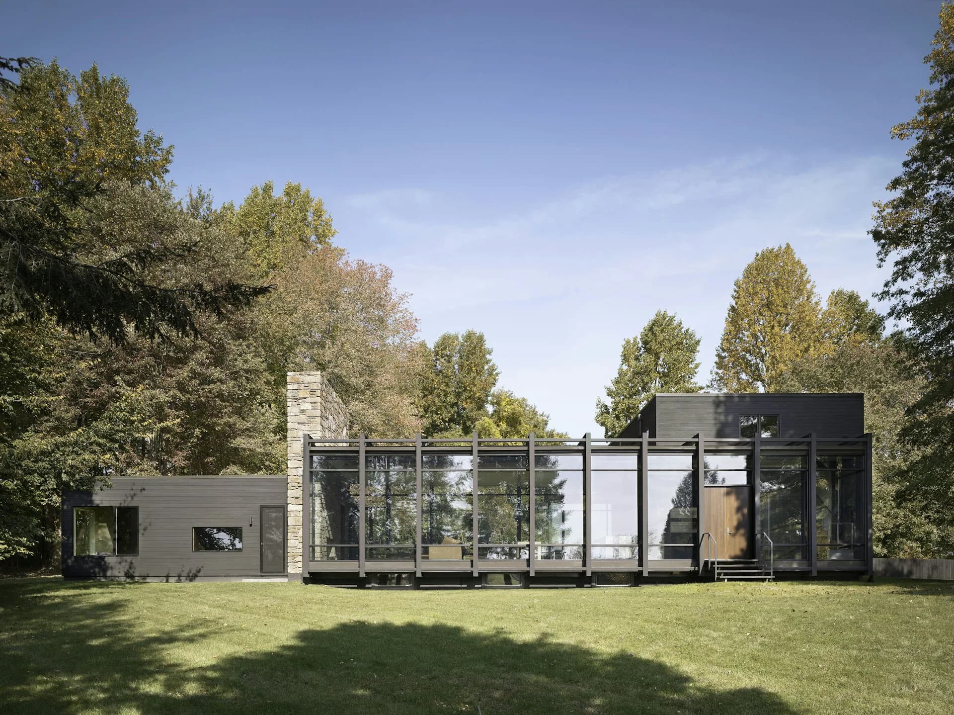 Modern residential architecture with black exterior cladding, extensive glazing, and stone chimney set in a verdant landscape by Koko Architecture + Design.