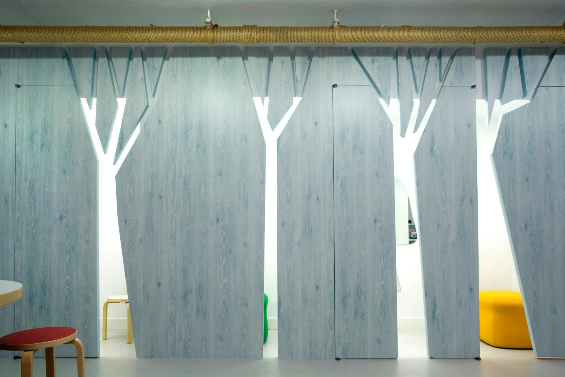 Modern interior space with floor-to-ceiling curtains featuring minimalist tree branch silhouettes in white against soft blue-gray fabric, accented with colorful seating elements by Koko Architecture + Design.