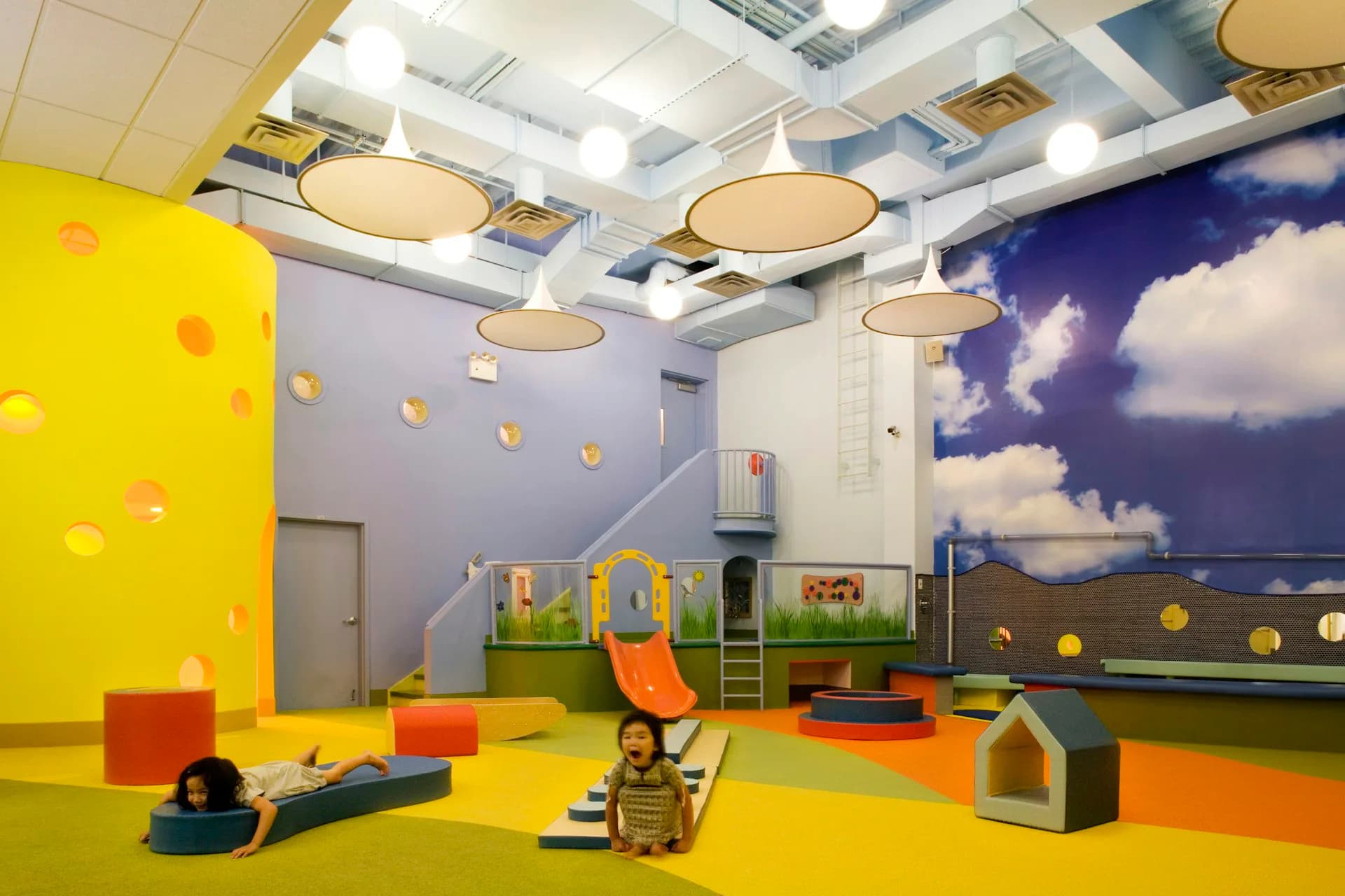 Colorful children's play space featuring bright yellow flooring, cloud-themed wall murals, suspended circular ceiling lights, and varied play structures in orange and green tones by Koko Architecture + Design.