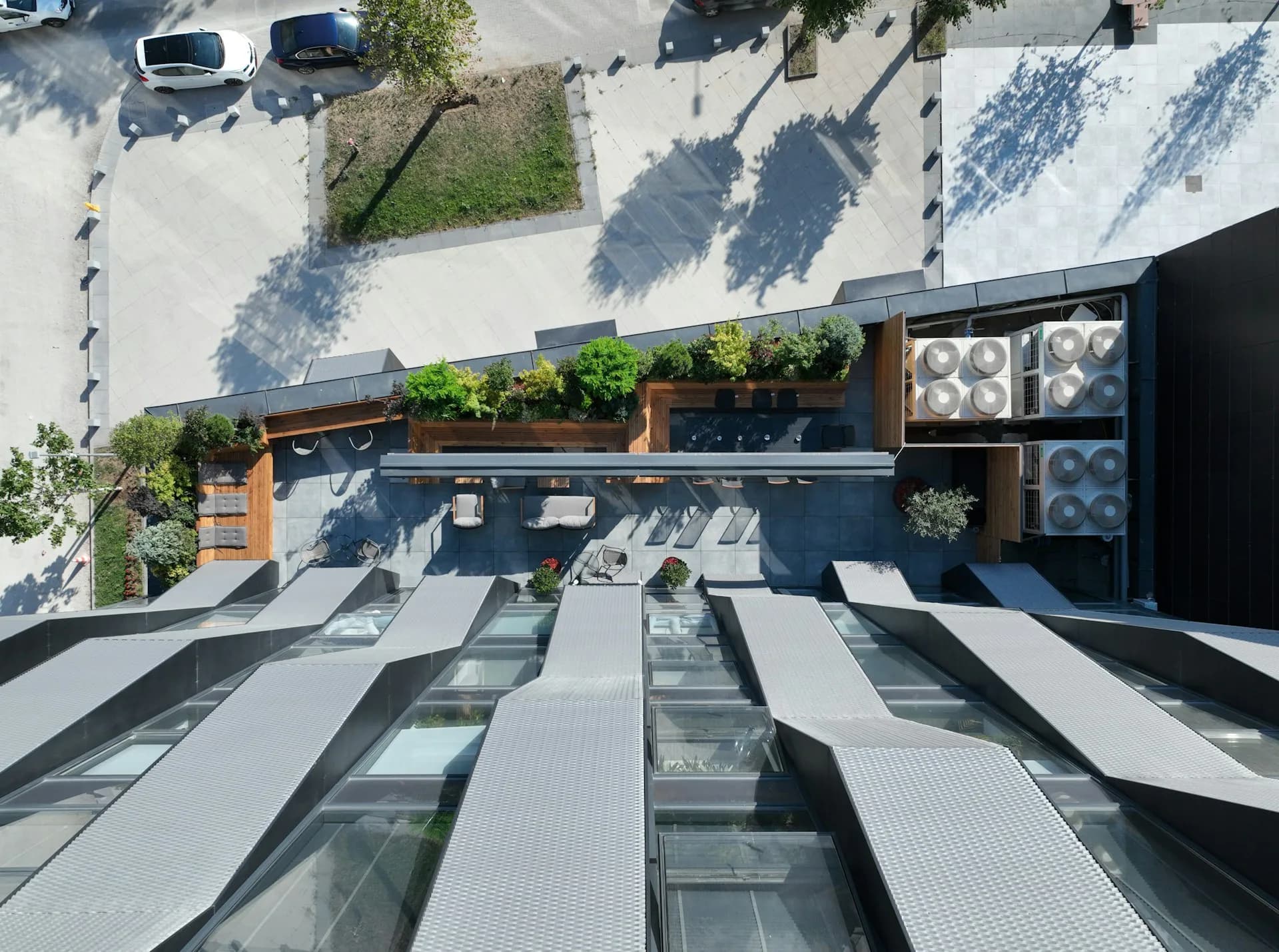Overhead view of a modern urban plaza with striped paving, green planters, seating areas, and parked cars, designed by Komoni Architecture.