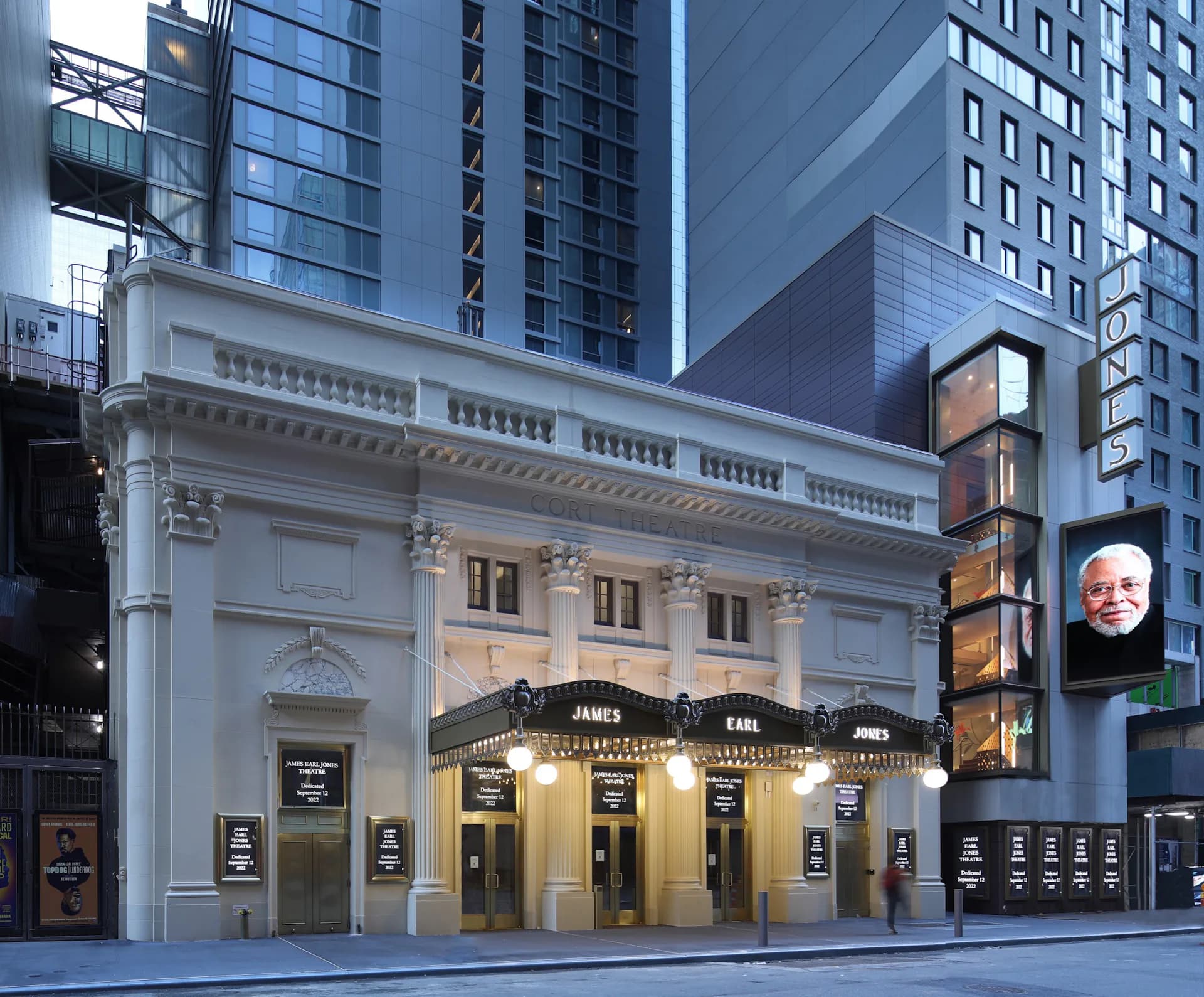 Historic neoclassical theater facade with white columns and ornamental details, illuminated at dusk among modern skyscrapers, by Kostow Greenwood Architects.