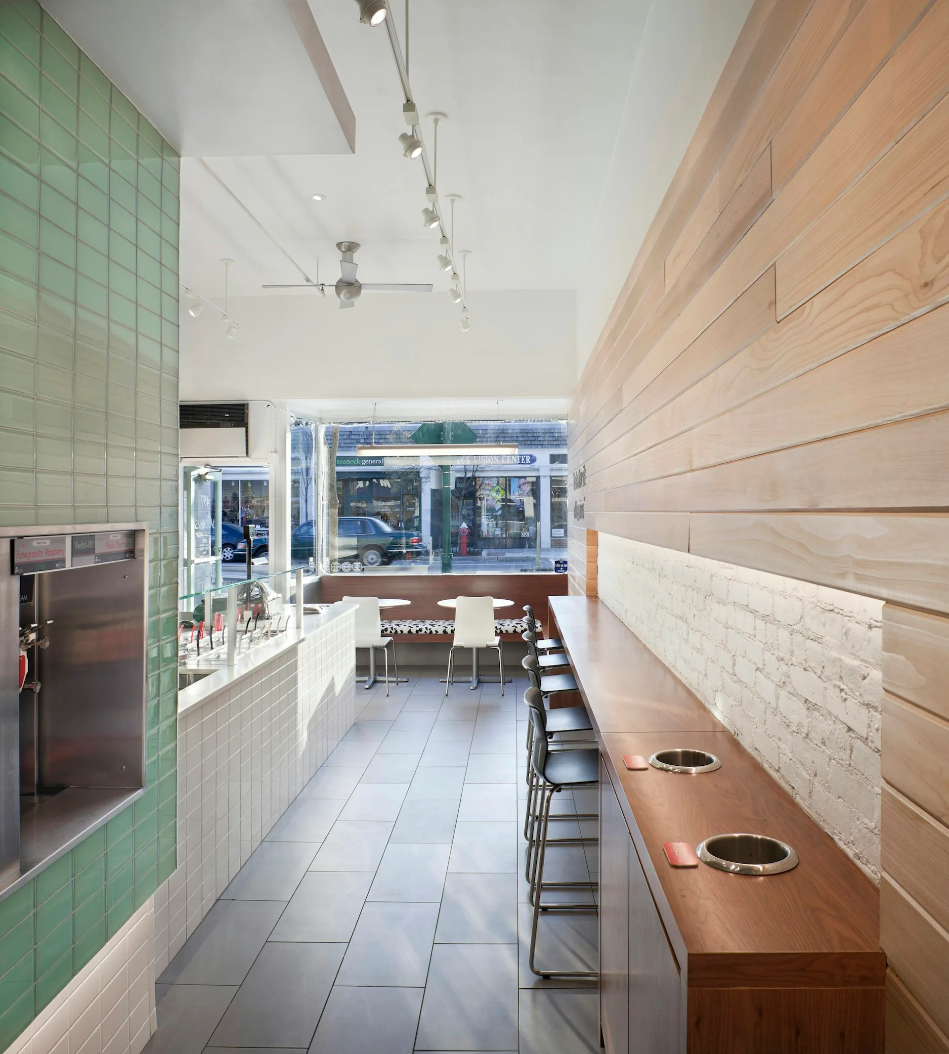 Long narrow restaurant or café interior with green tile and white brick walls, wood paneling on right side, counter seating along left, and large storefront windows at end by Krajewski Architect PLLC.