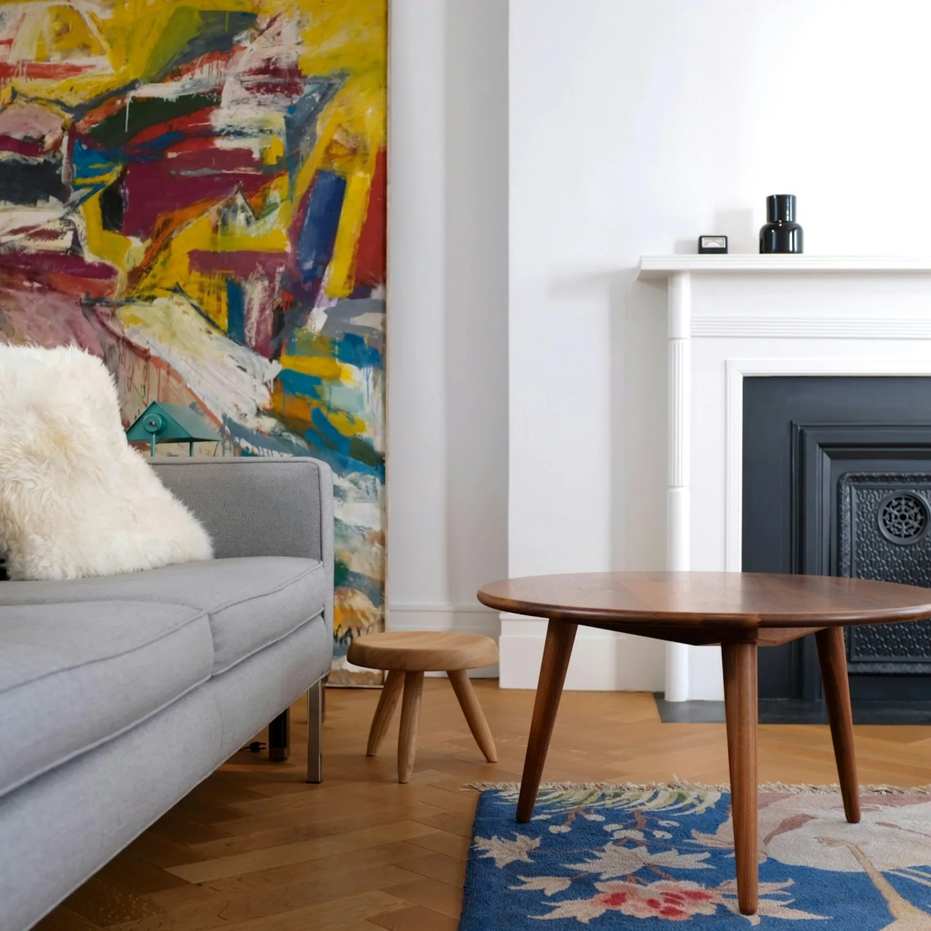 Modern living room with gray sofa, wooden mid-century coffee table, colorful abstract artwork, geometric blue rug, and fireplace by Krajewski Architect PLLC.