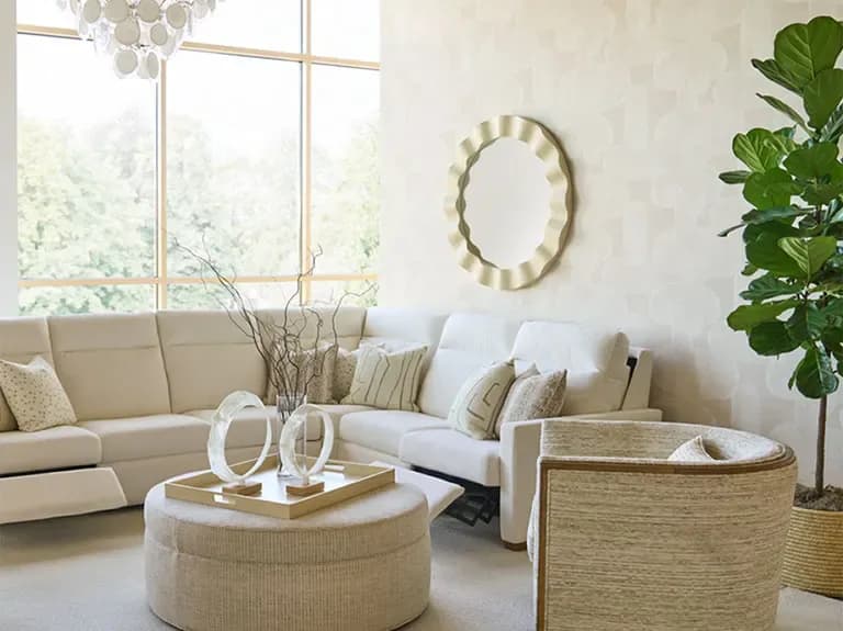 Neutral-toned living room with beige sectional, round woven ottoman, curved mirror, and potted plants creating a light, airy aesthetic by Kravet Inc.