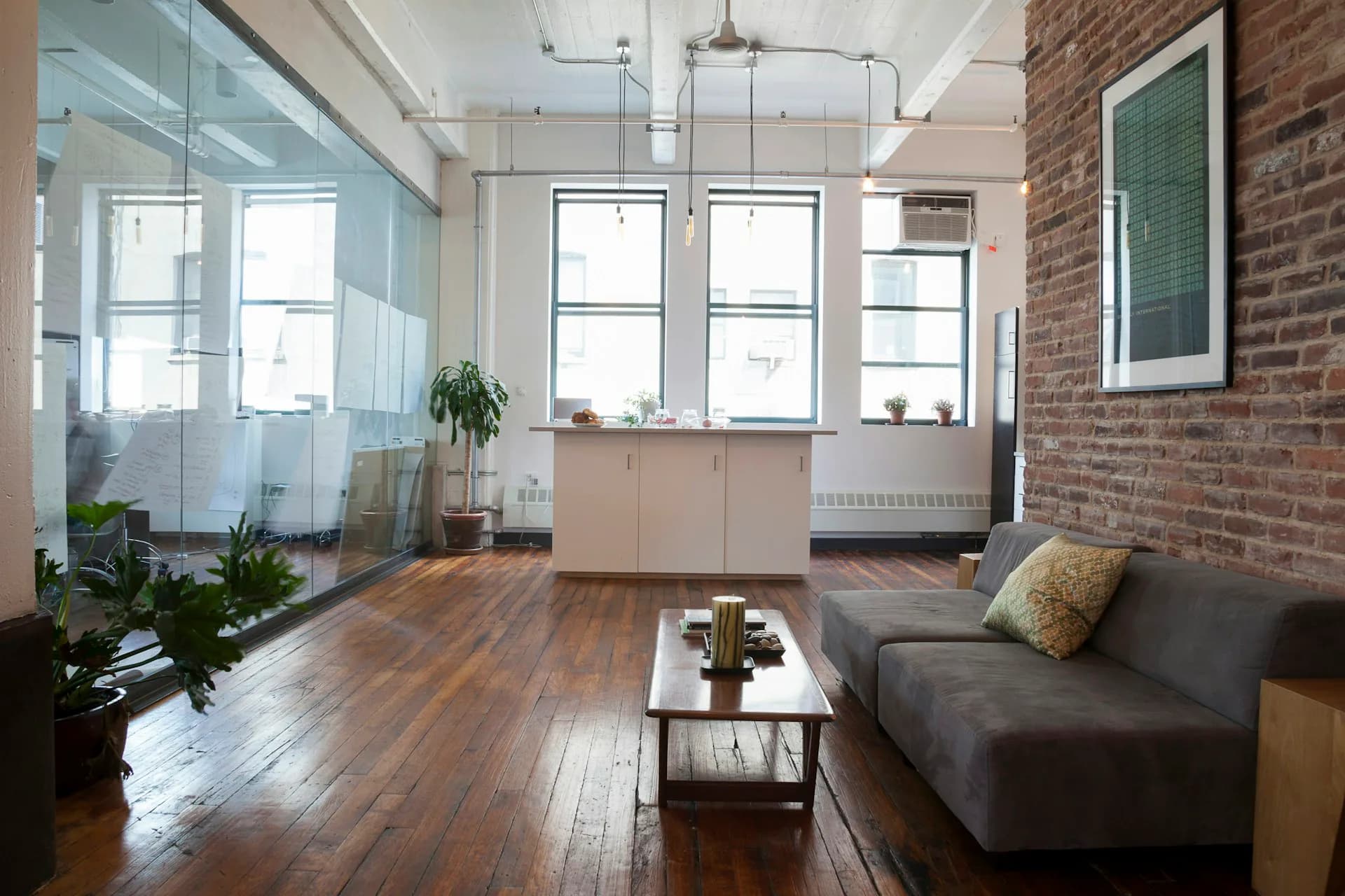 Modern loft interior featuring exposed brick walls, wood flooring, large windows, gray seating, and glass-partitioned office space by +LAB Architect PLLC.