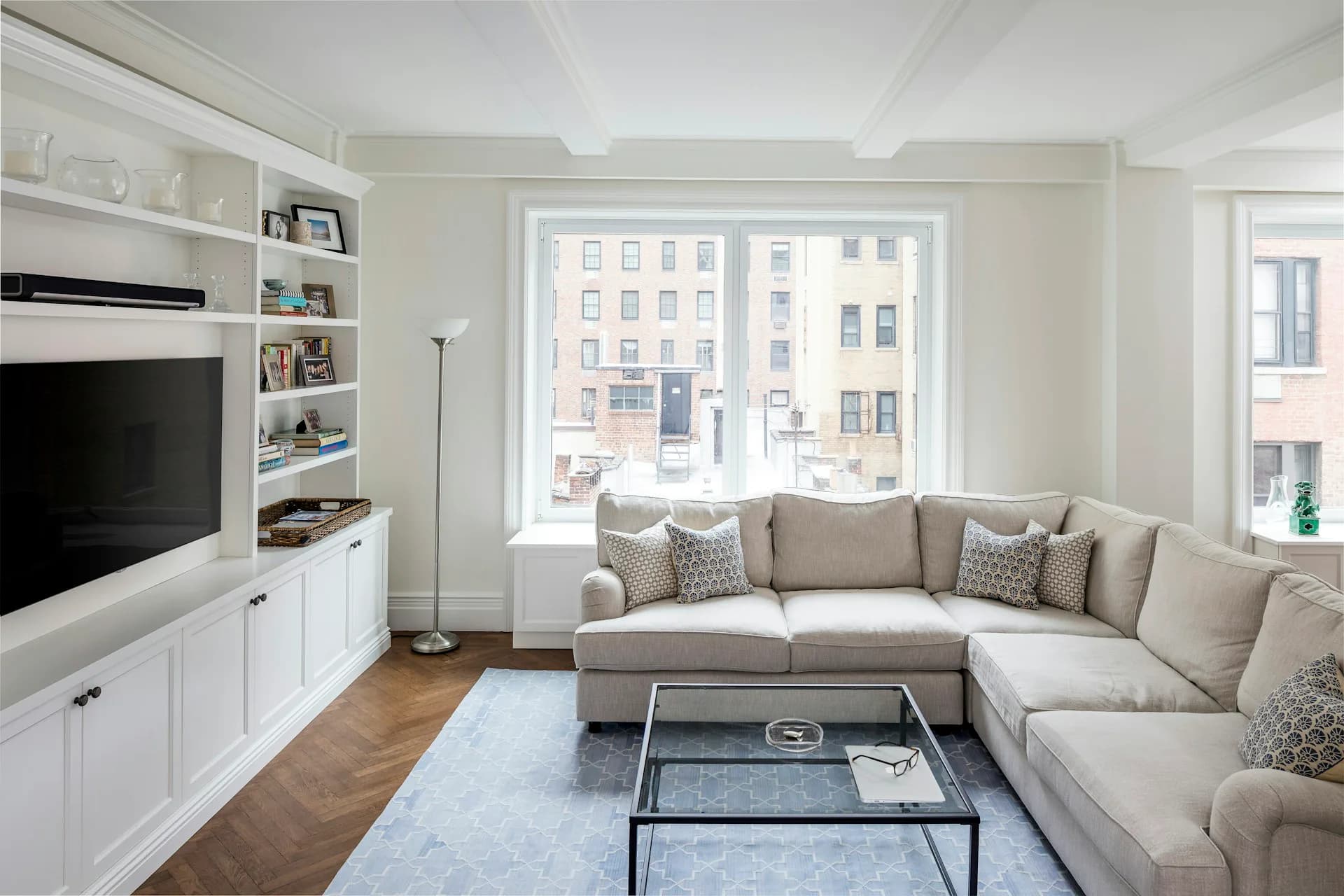 Bright living room with beige L-shaped sectional, white built-in cabinetry, hardwood floors, and large windows overlooking urban buildings by +LAB Architect PLLC.