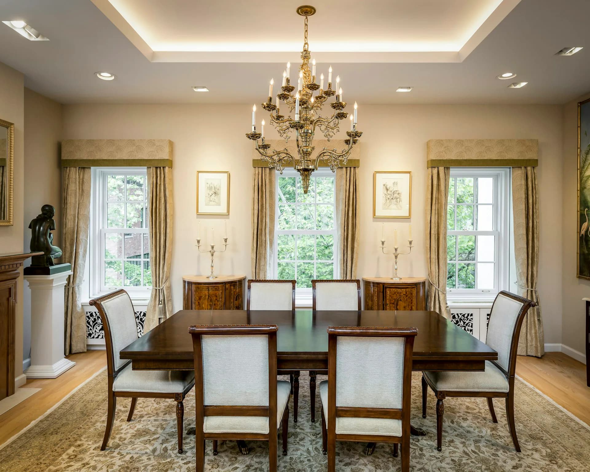 Formal dining room with wood table, upholstered chairs, ornate brass chandelier, cream walls, and garden views designed by +LAB Architect PLLC.
