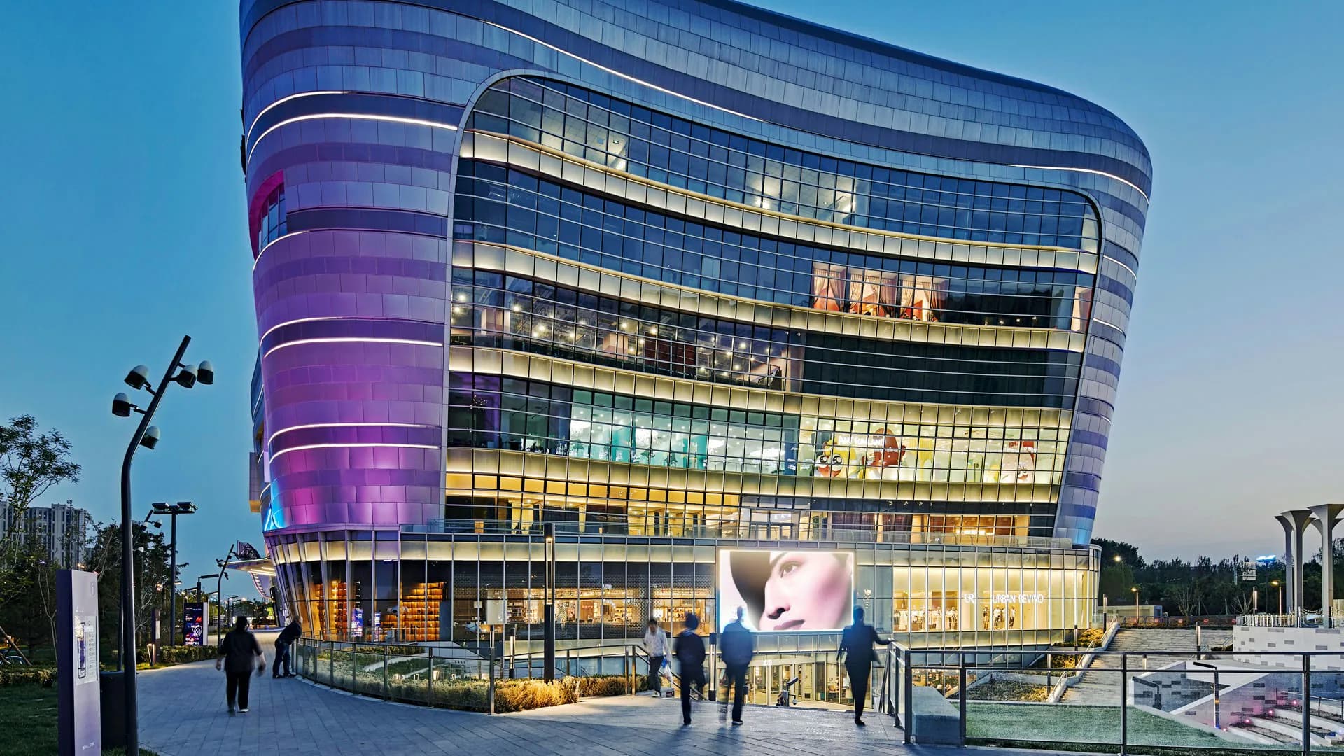 Curved modern building with illuminated purple and gold facade, glass curtain wall with horizontal striping, plaza with people and street lamps at dusk by LAGUARDA.LOW ARCHITECTS.