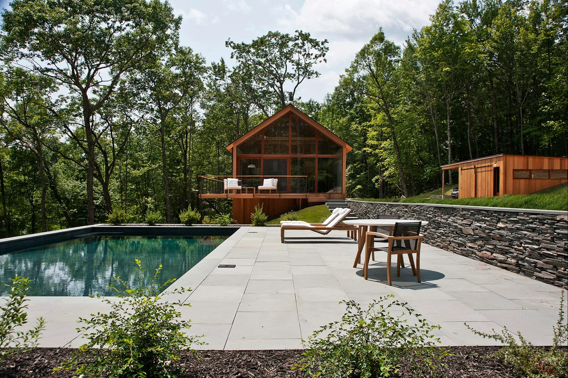 Modern residential pool deck with concrete paving, lounge furniture, and wooden architecture nestled in a forested setting by Lang Architecture.