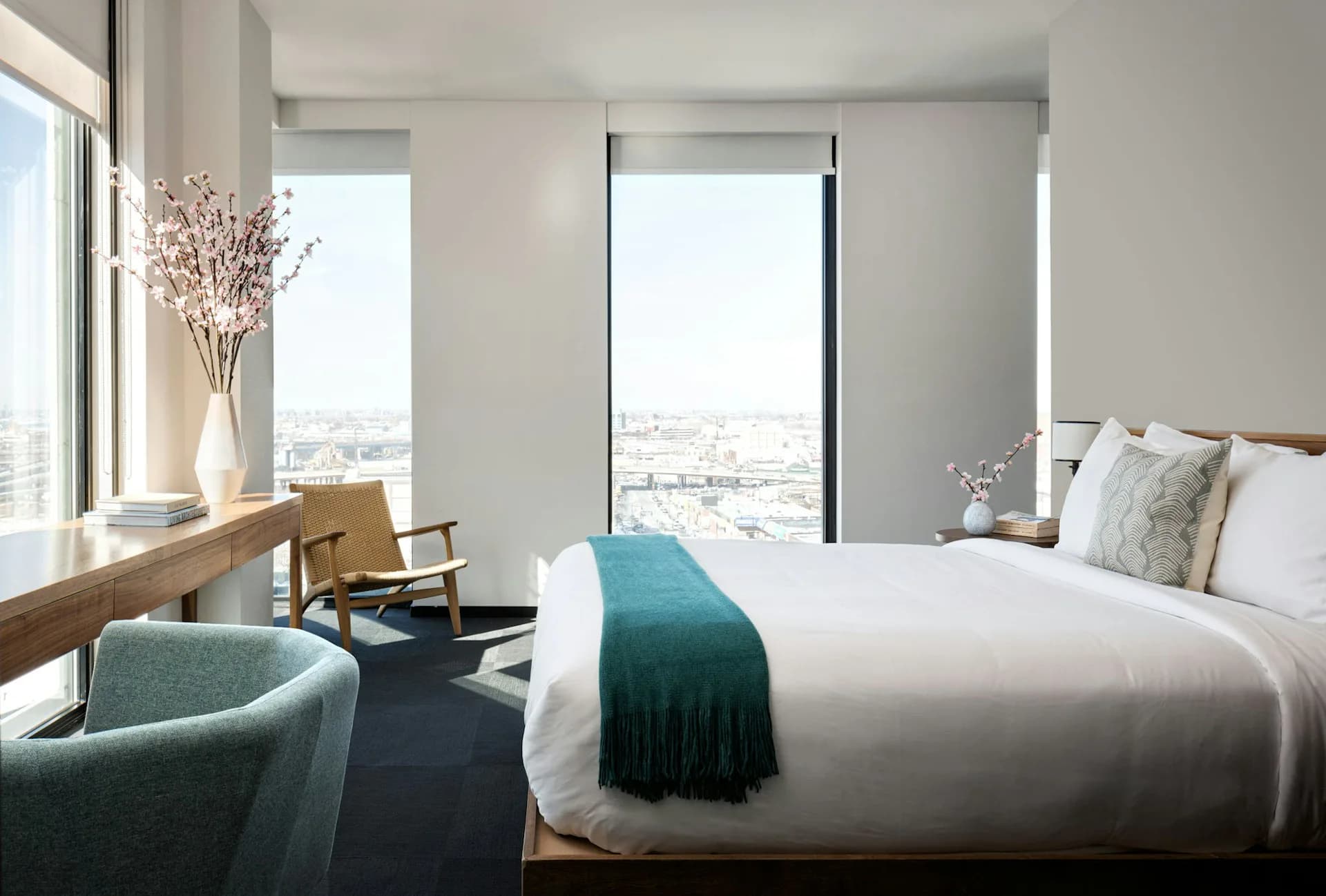 Modern hotel bedroom with floor-to-ceiling windows overlooking cityscape, white bedding, teal accent chair, natural wood desk, and minimalist palette by Lang Architecture.