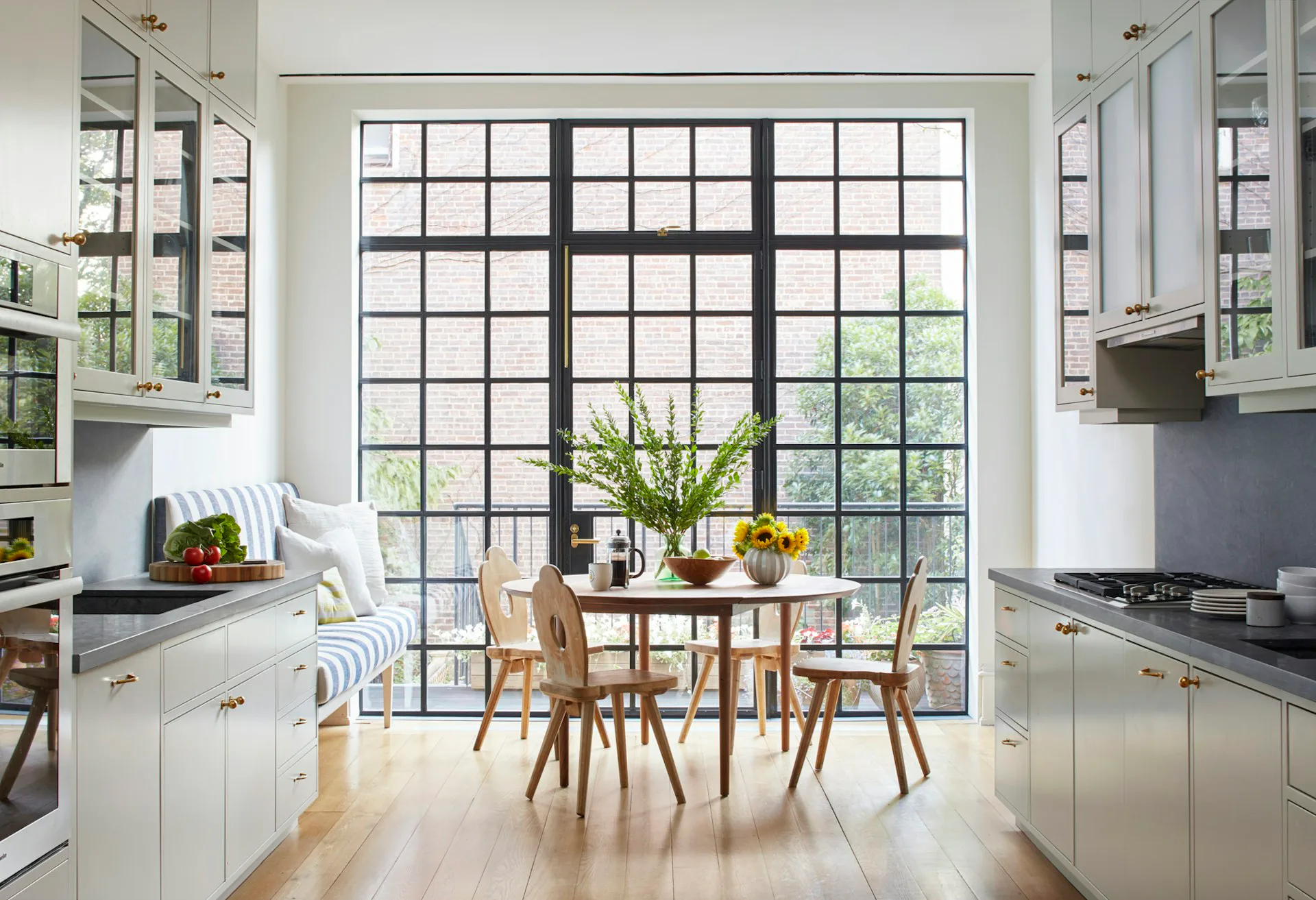 Bright galley kitchen with white cabinetry flanking large gridded windows overlooking a dining area with wooden chairs and table by Lang Architecture.