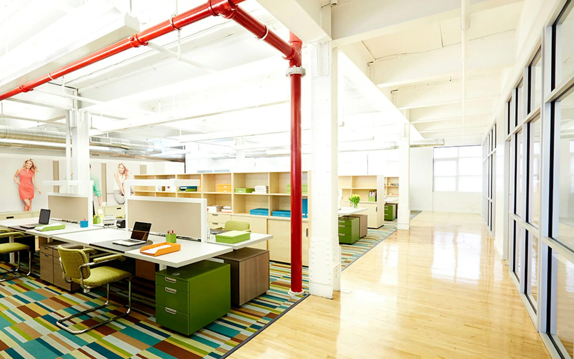 Bright, open-plan office workspace featuring a red structural column, green storage cabinetry, colorful striped flooring, wooden desks, and natural lighting by Larson Architecture Works pllc.