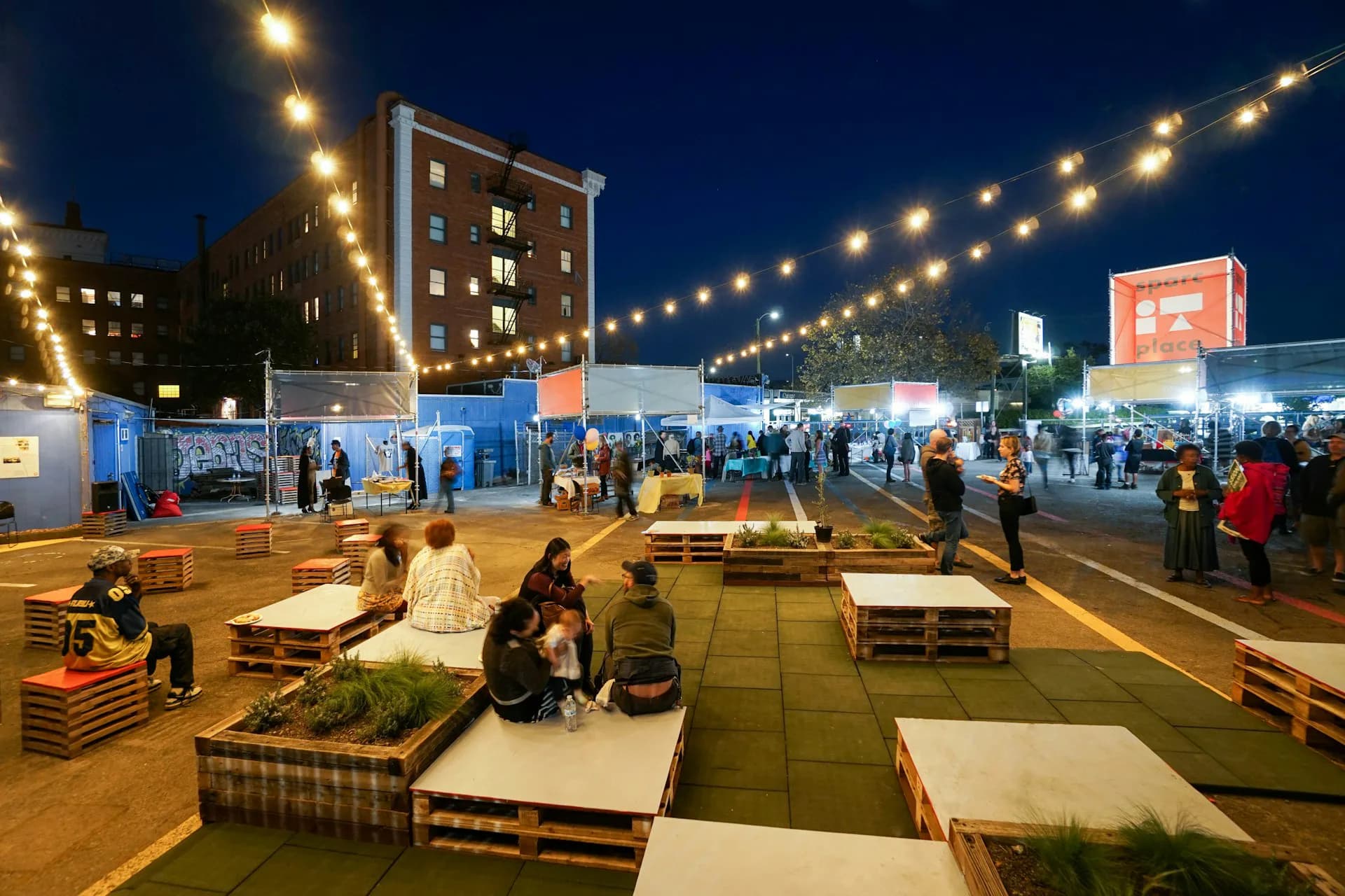 Evening view of an urban plaza with wooden pallet seating arrangements, string lighting, and blue pavilions, designed by Leibin Architecture.