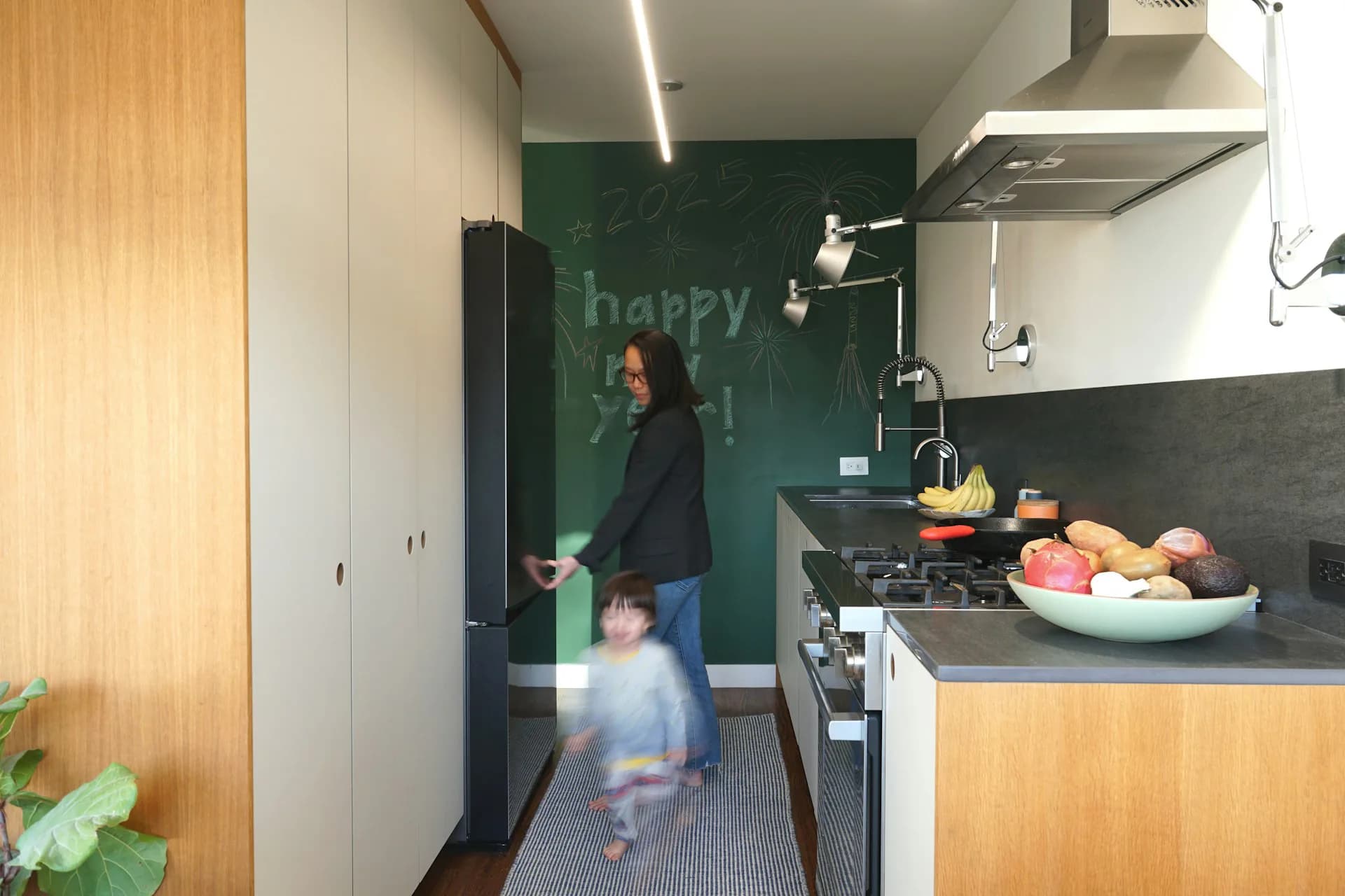 Modern kitchen with warm wood cabinetry, black countertops, and a green chalkboard accent wall, featuring open shelving and stainless steel appliances by Leibin Architecture.