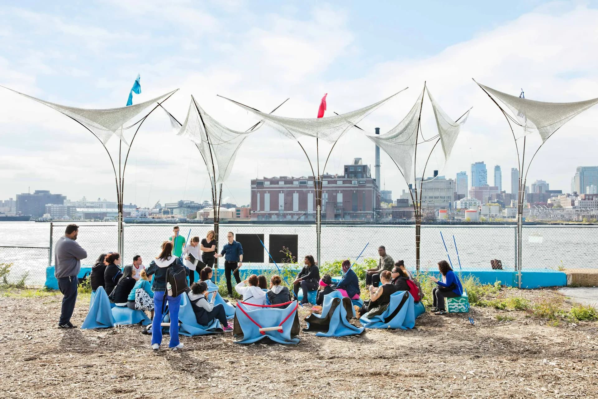 Waterfront pavilion with tensile fabric canopies sheltering a gathered group on sandy beach, Manhattan skyline beyond, by Leroy Street Studio.
