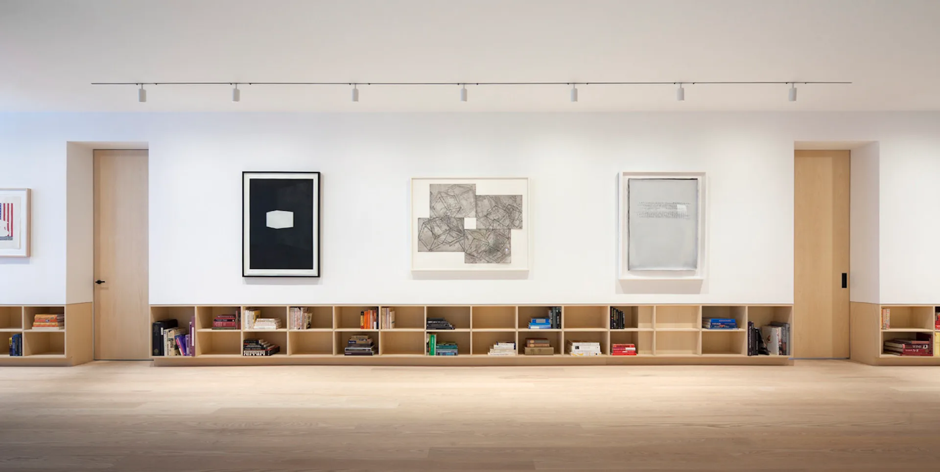 Minimalist gallery interior with continuous wooden shelving system, white walls, track lighting, and framed artworks by LEVENBETTS.
