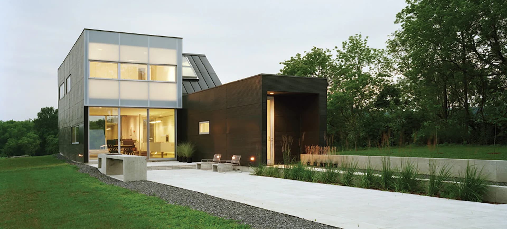 Modern residential home with illuminated glass and dark wood exterior set in green landscape by LEVENBETTS.
