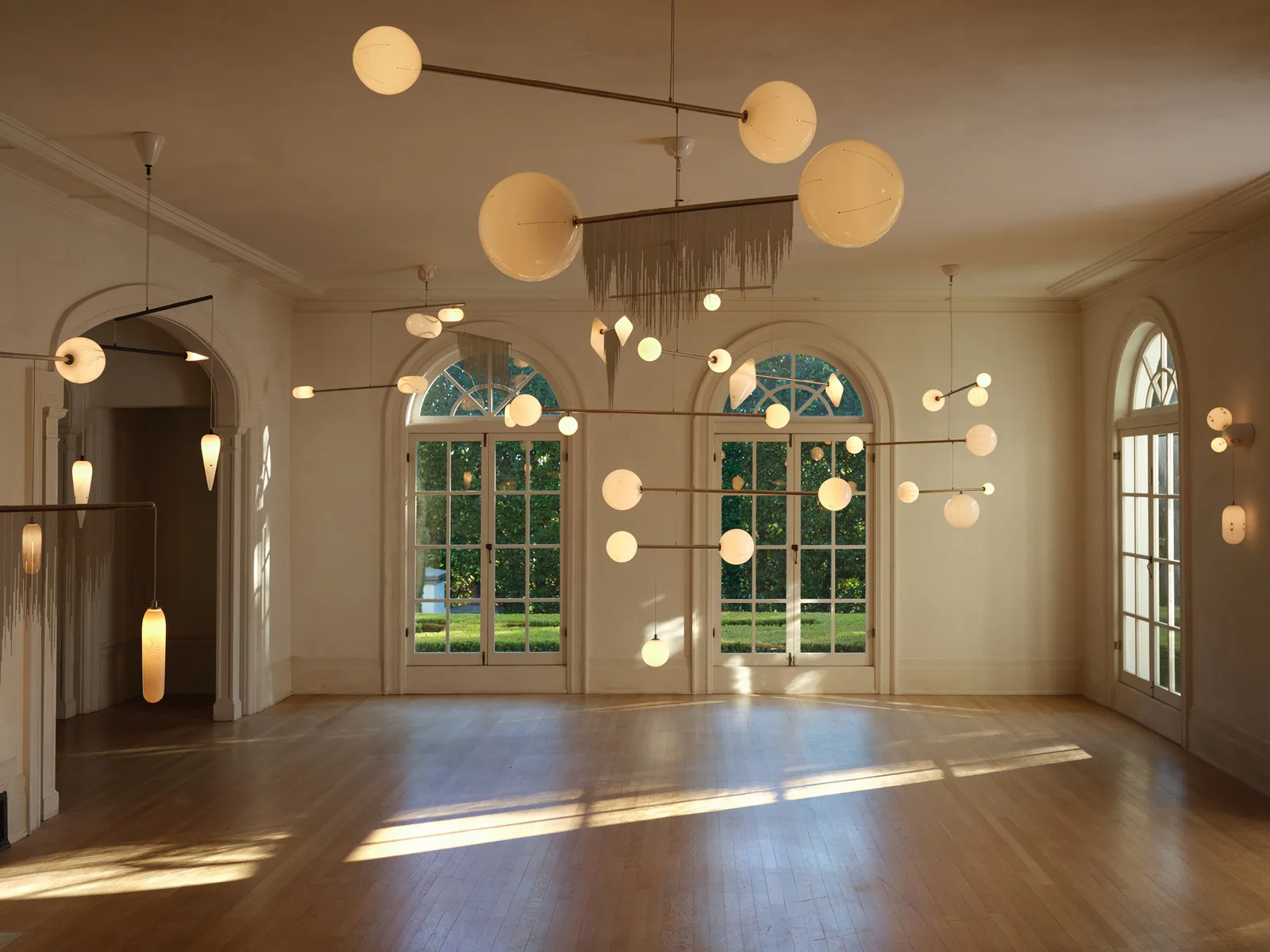 Spacious gallery with arched windows, wooden floors, and suspended spherical lighting fixtures in warm tones by Lindsey Adelman Studio.