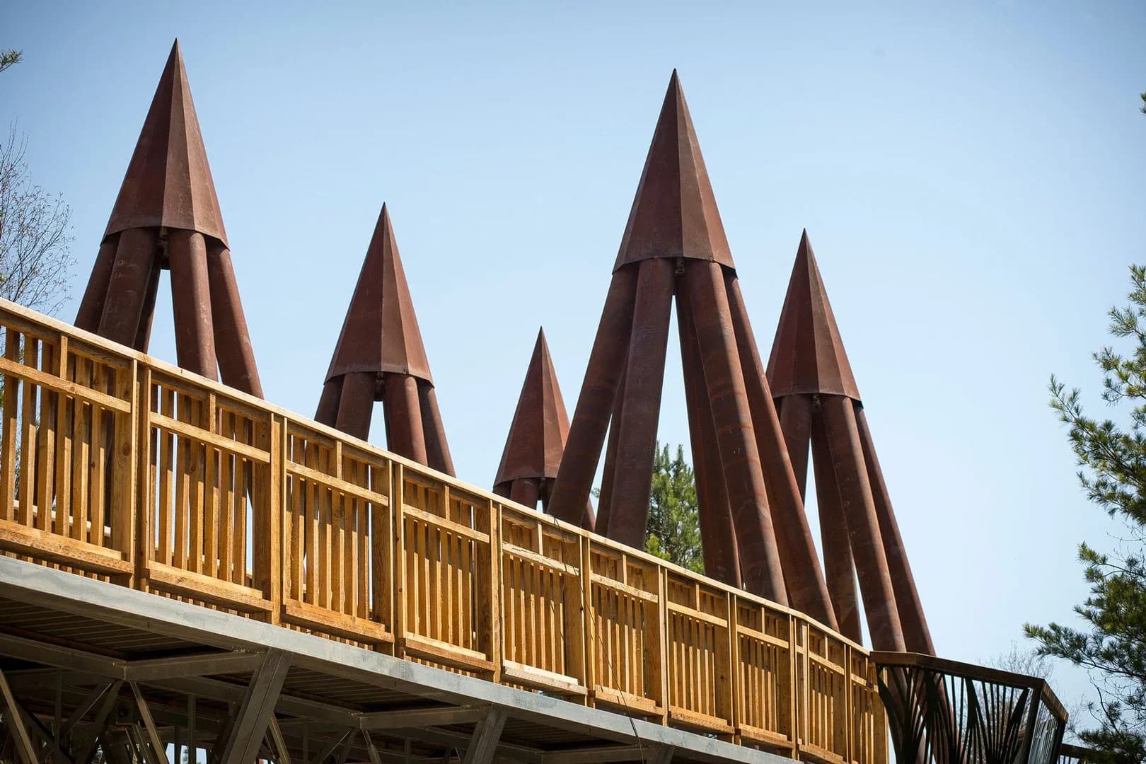 Outdoor pedestrian bridge with yellow wooden railings and striking rust-brown conical metal spires against a clear sky, designed by Linearscape.