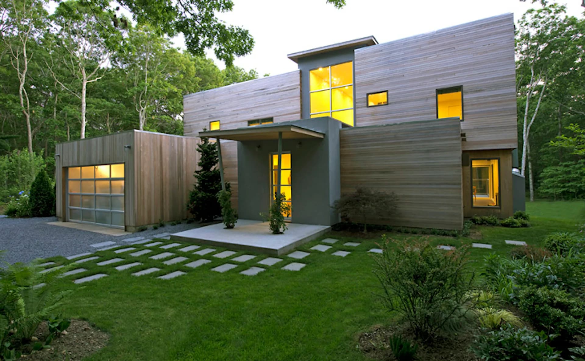 Modern residential home with dark fiber cement cladding, large yellow-lit windows, and integrated garage, set on manicured lawn by Lisa Dubin Architect.